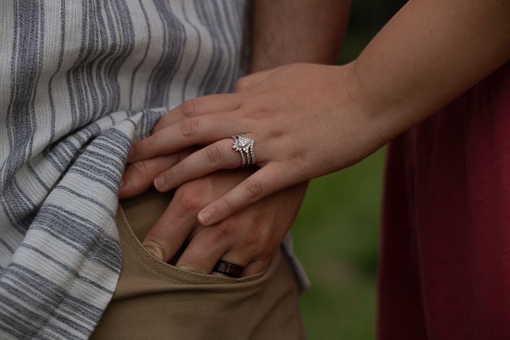 Close-up of a woman's hand with a diamond engagement ring resting on a man's hand outdoors.
