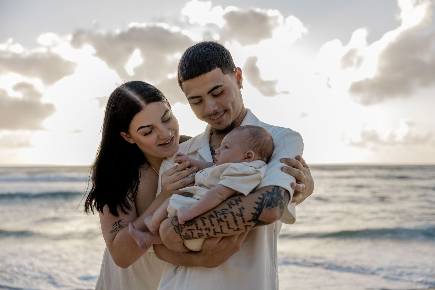 Happy young parents smiling and holding newborn baby together on beach at golden sunset.
