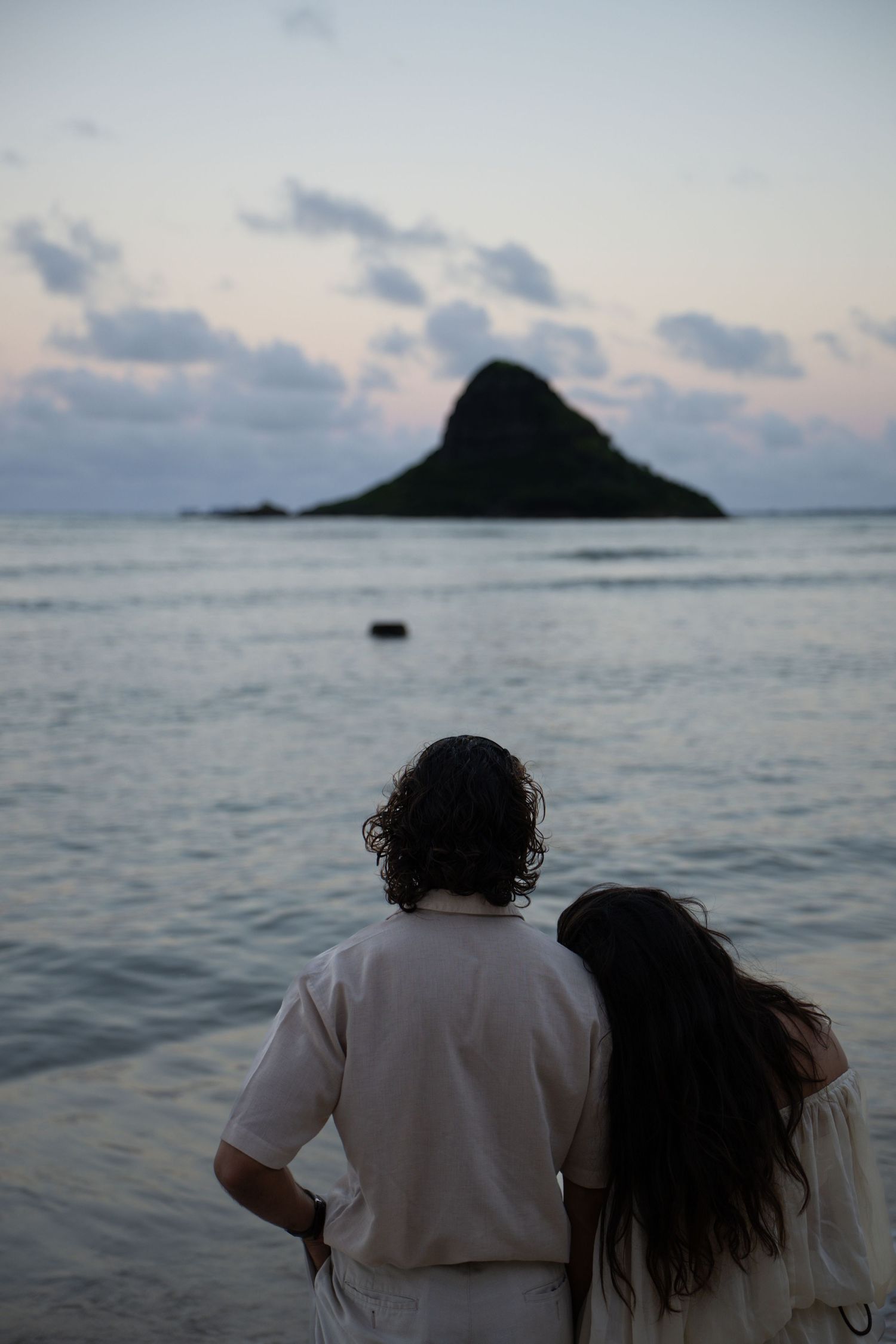 Couple standing back to camera viewing Chinaman Hat island at dusk in Hawaii.