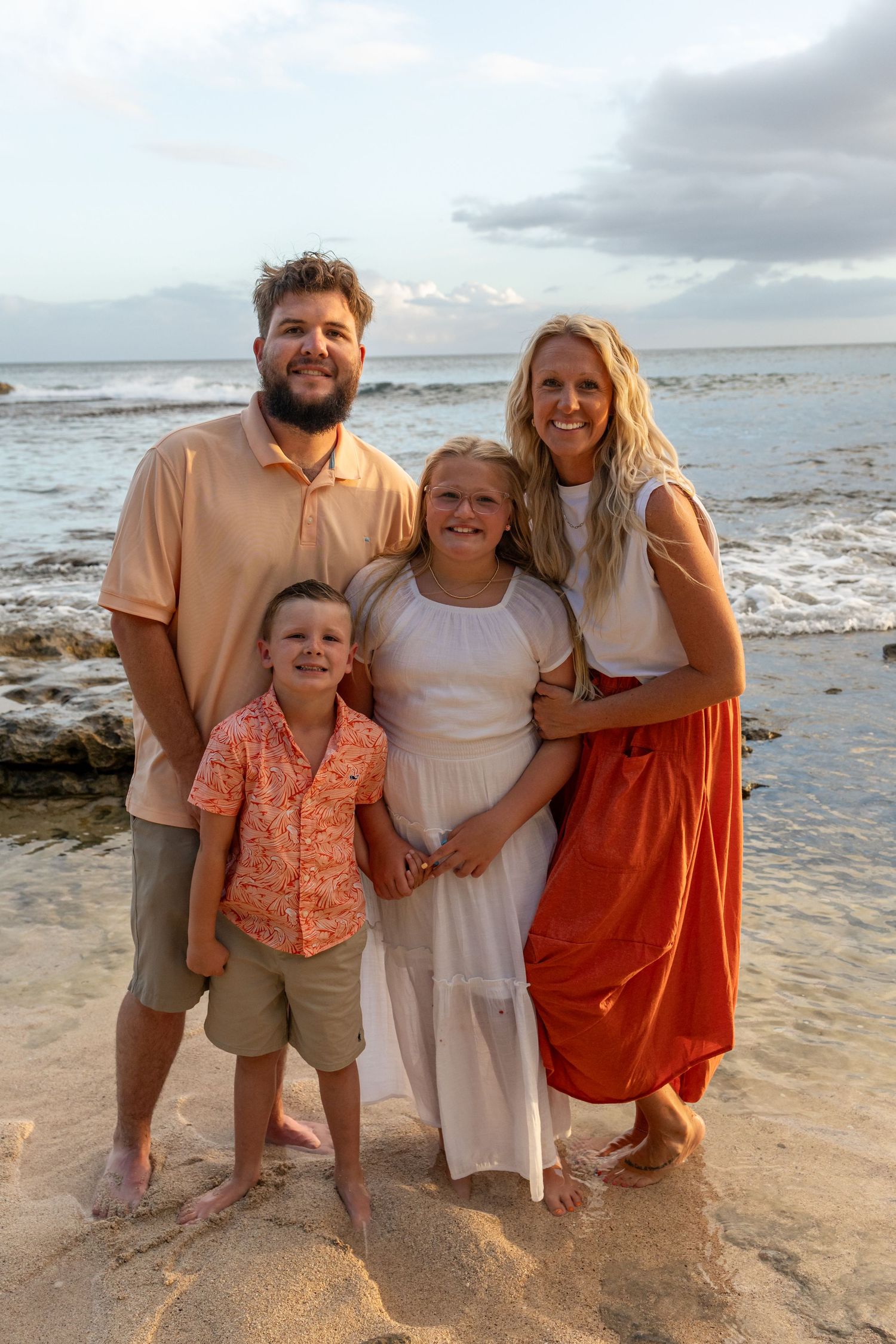 Family of four smiling together on a sandy beach at sunset, wearing coordinated outfits in warm tones.