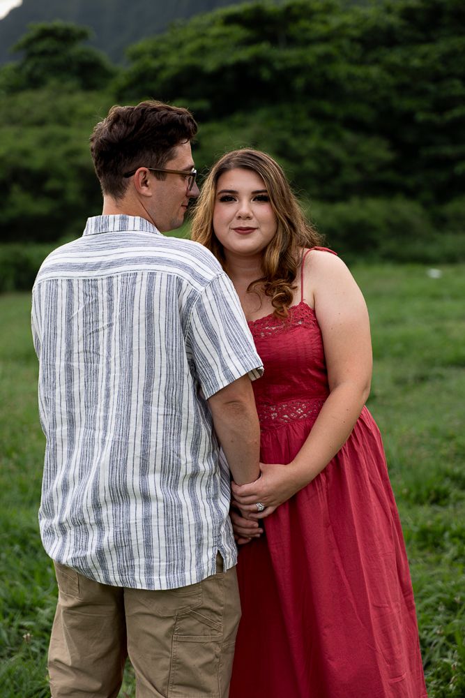Couple holding hands in green field, woman in red lace dress smiling at man in striped shirt.