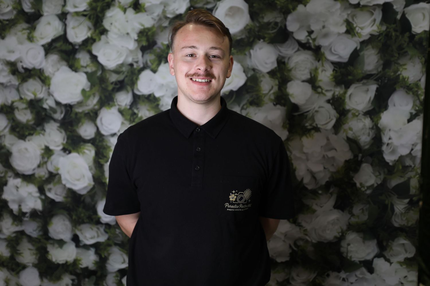 Young man smiling in black polo shirt against white floral wall backdrop.