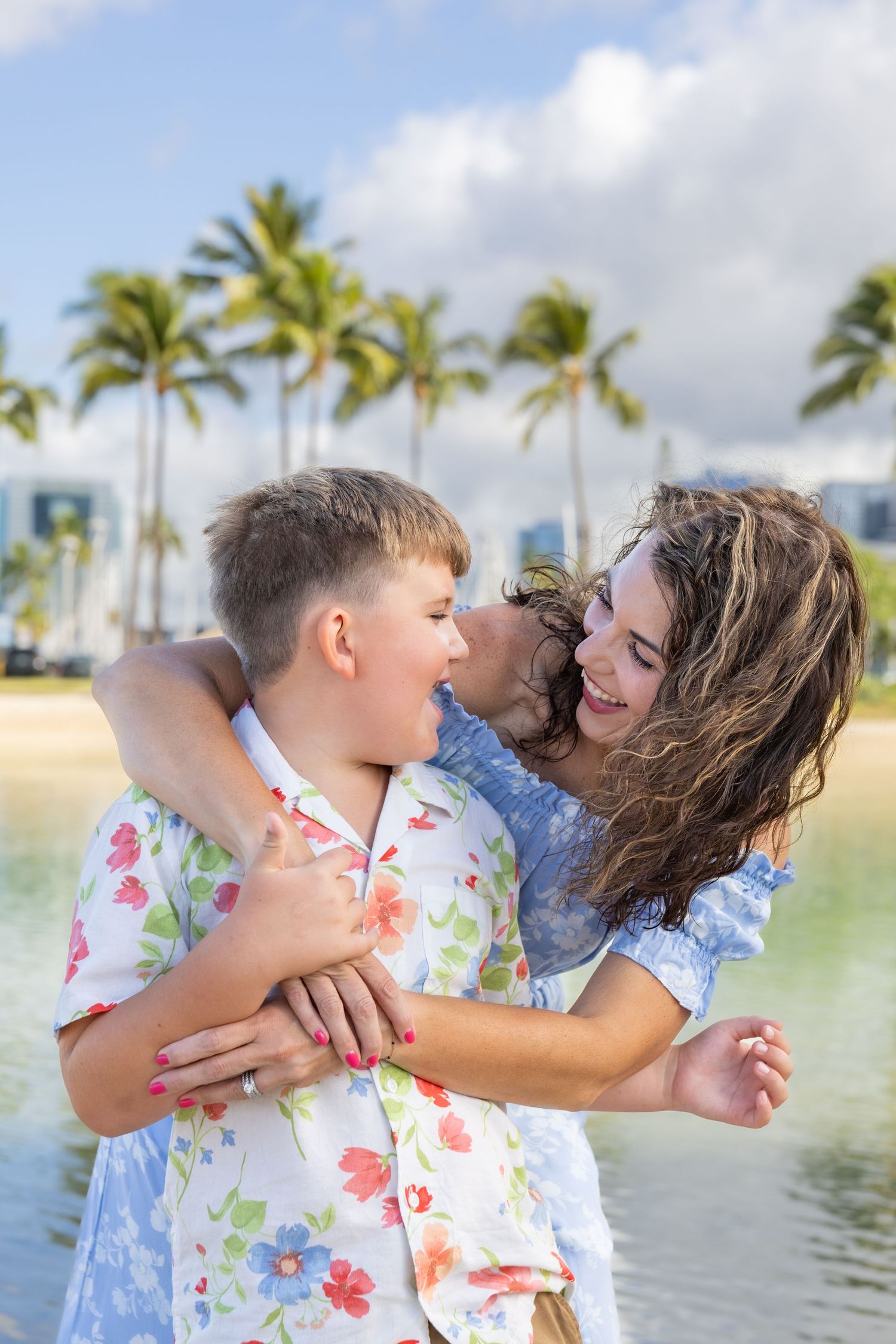A joyful mother hugging her two children on a tropical beach with palm trees and blue sky.