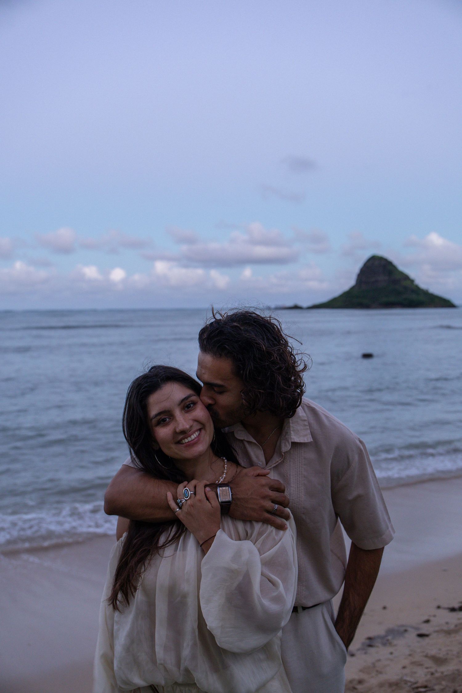 Couple embracing on a Hawaii beach at dusk with iconic Chinaman's Hat island rock formation in background.