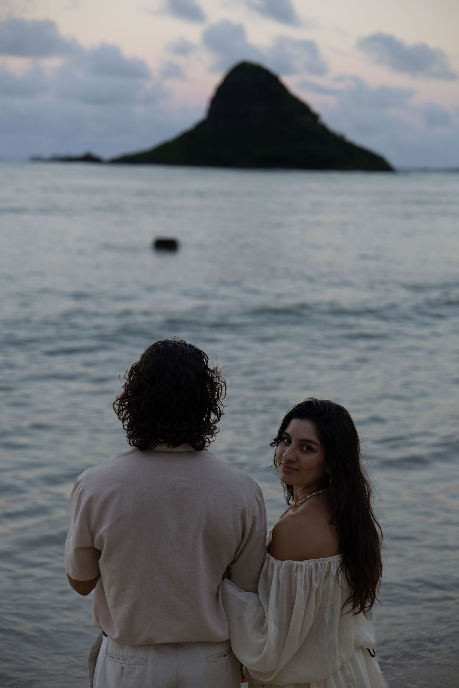 Couple standing on beach at sunset with iconic Chinaman Hat island in background.