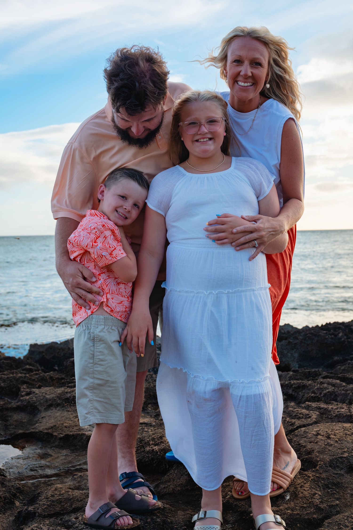 Family of four posing together on rocky shoreline with ocean and cloudy sky backdrop.