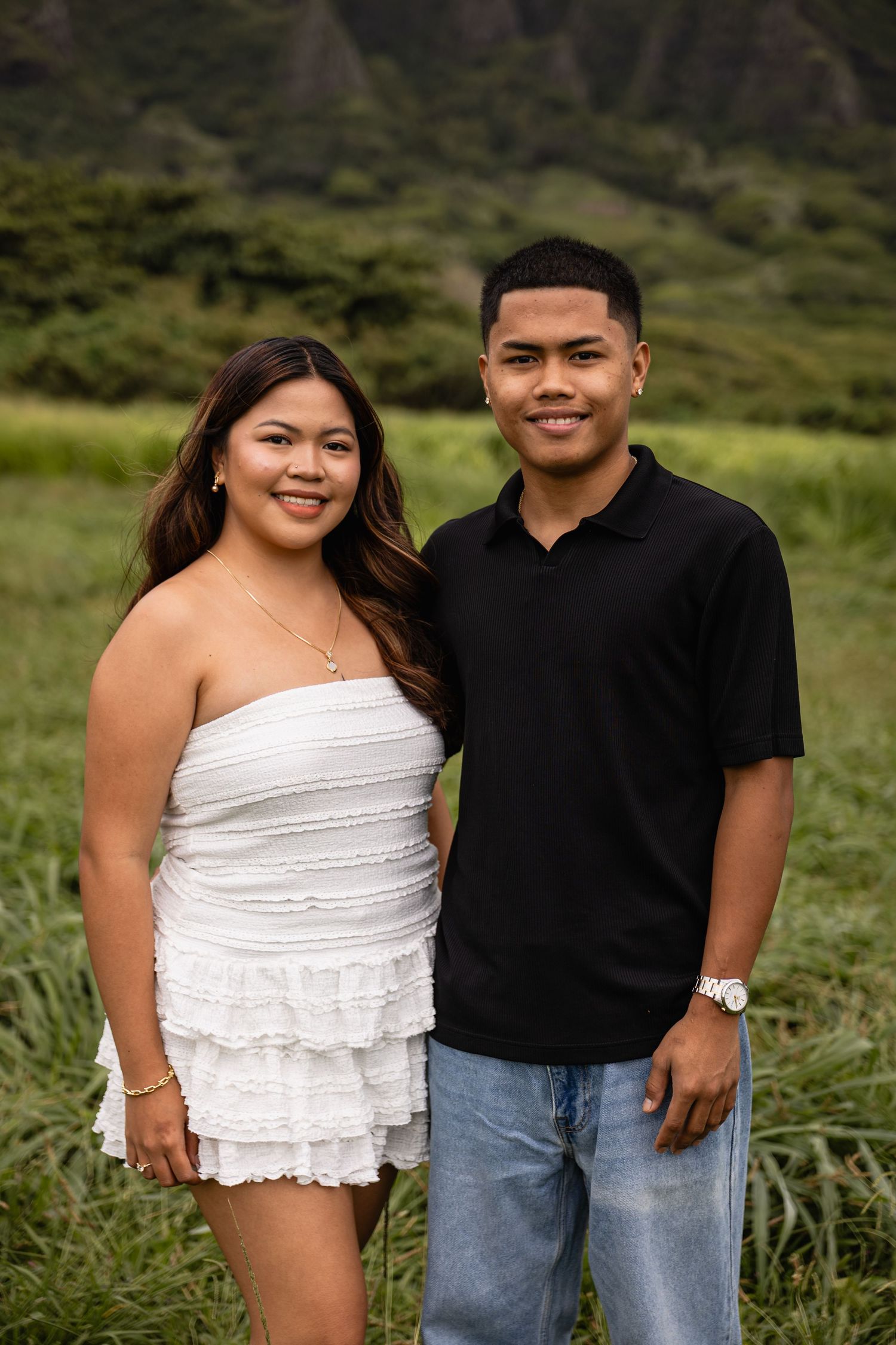 A young couple poses outdoors in Hawaii, woman in white dress and man in black polo shirt, with lush green mountains behind.