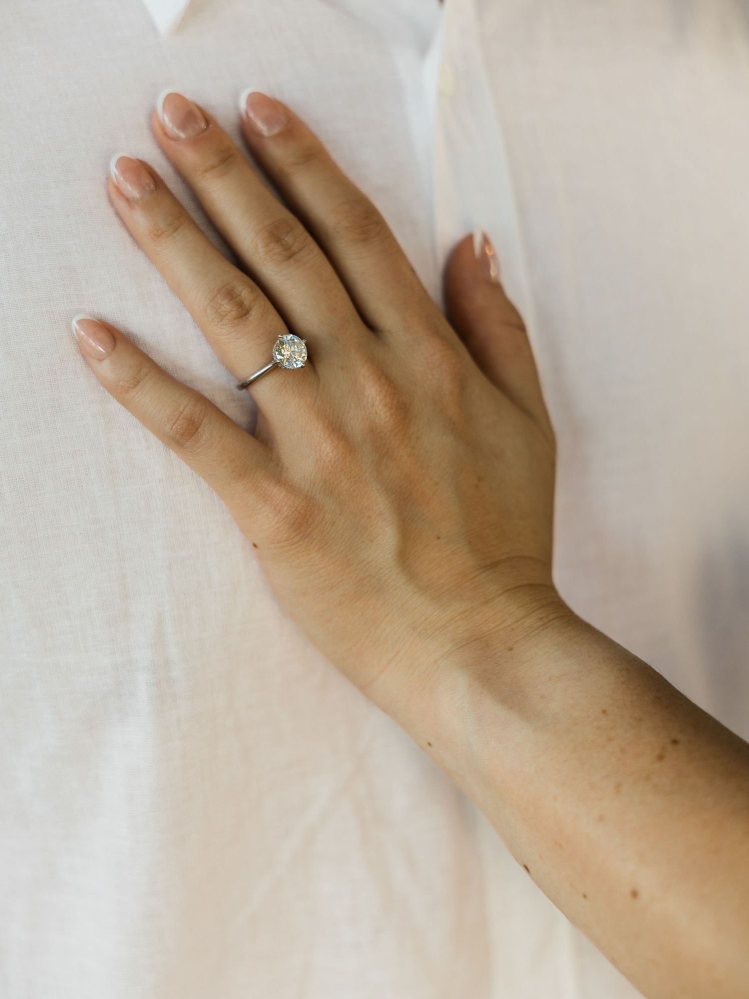 Close-up of woman's hand wearing elegant solitaire diamond engagement ring on white fabric.