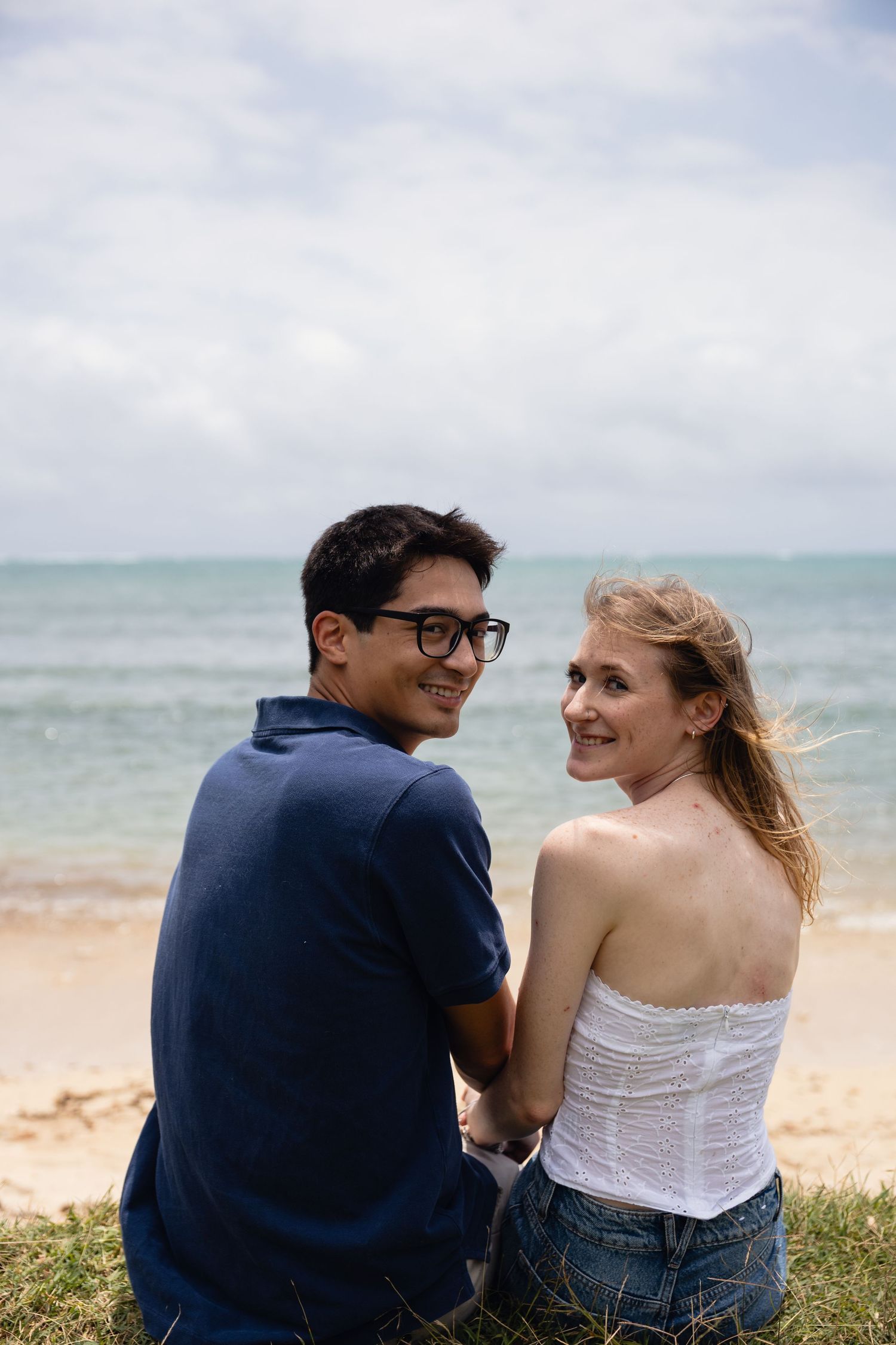 A smiling couple sits on a grassy beach, holding hands with the ocean in the background.