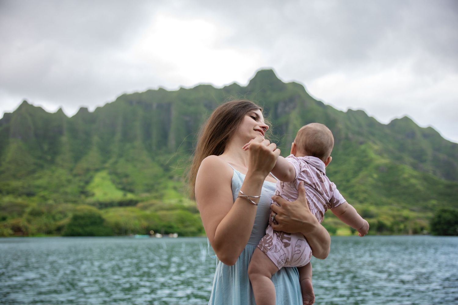 Mother holding baby girl by scenic Hawaii mountain lake in blue dress.
