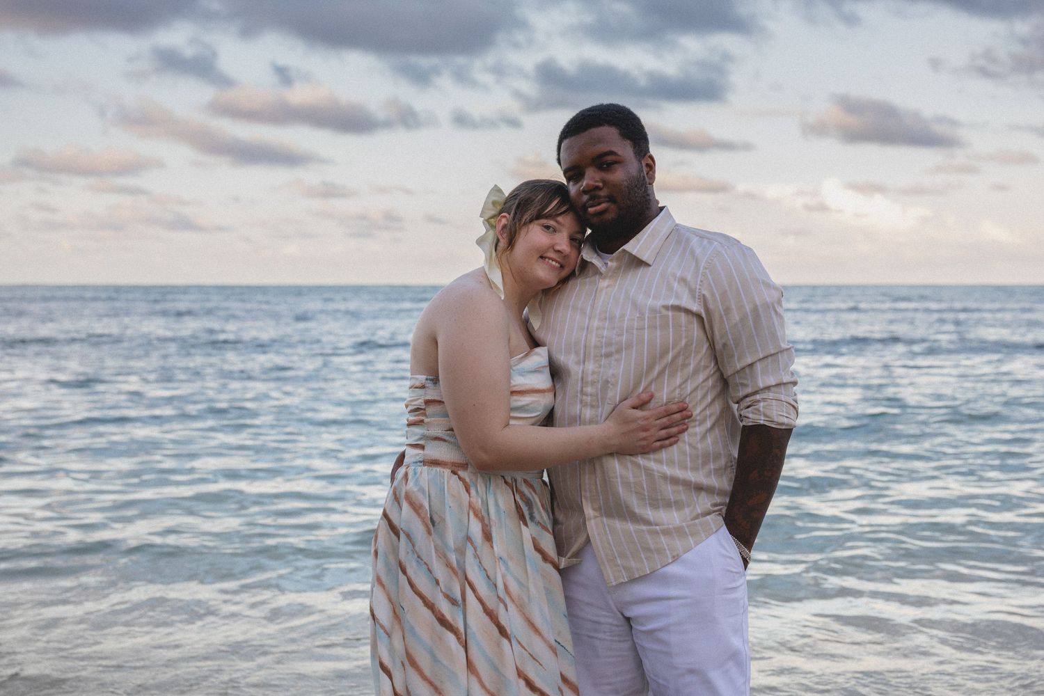 Couple embracing on a beach at sunset, woman in striped dress and man in white pants, with ocean waves in the background.