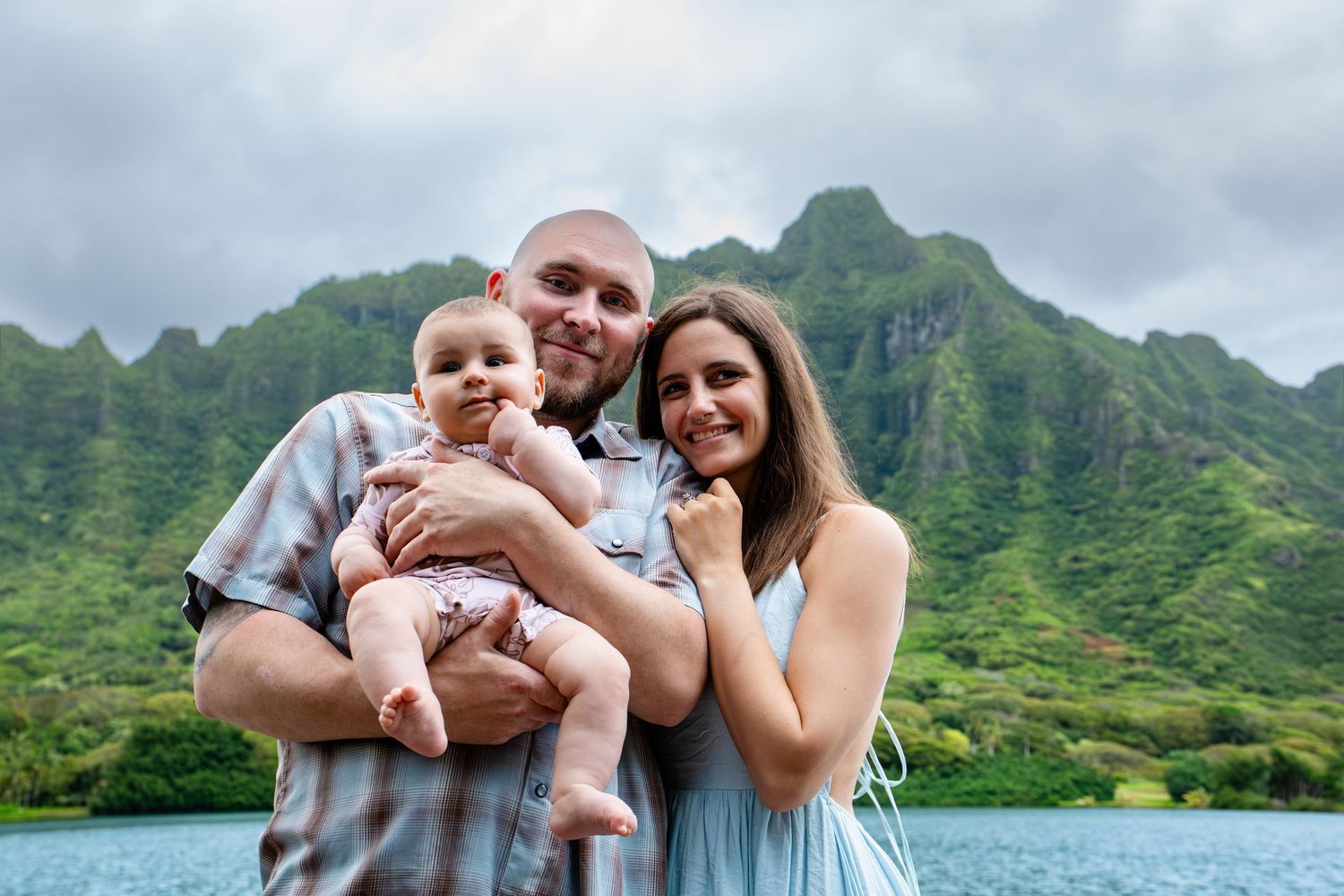 Smiling family holding baby in front of lush green Hawaii mountain lake.