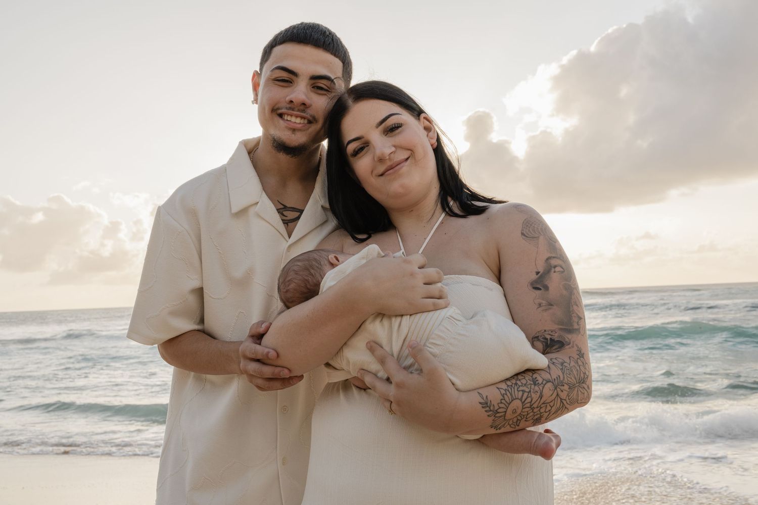 Young couple with newborn baby posing on beach at sunset, woman with tattoos in white dress.