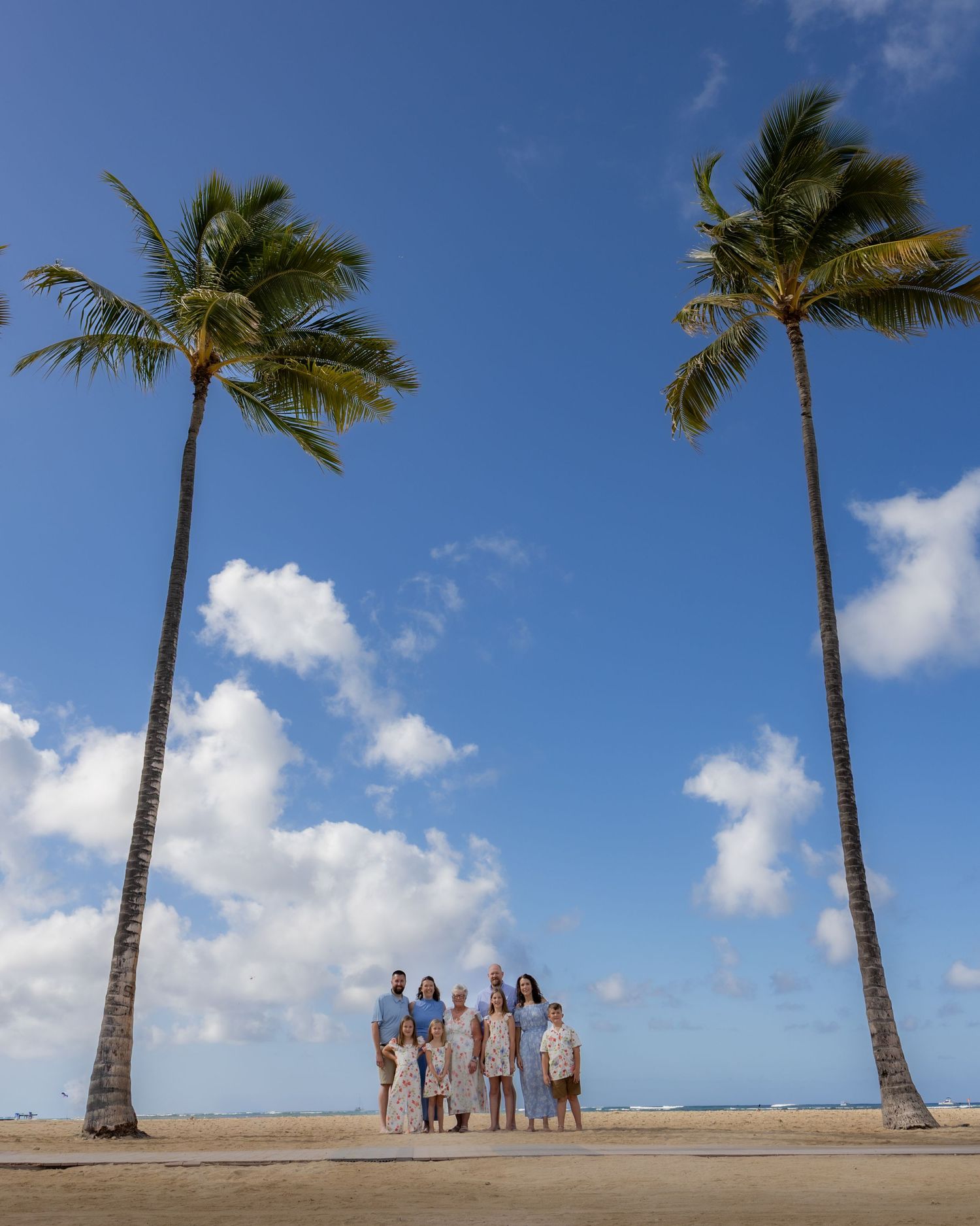 Family group posing between two tall palm trees on a sunny tropical beach with blue sky and white clouds.