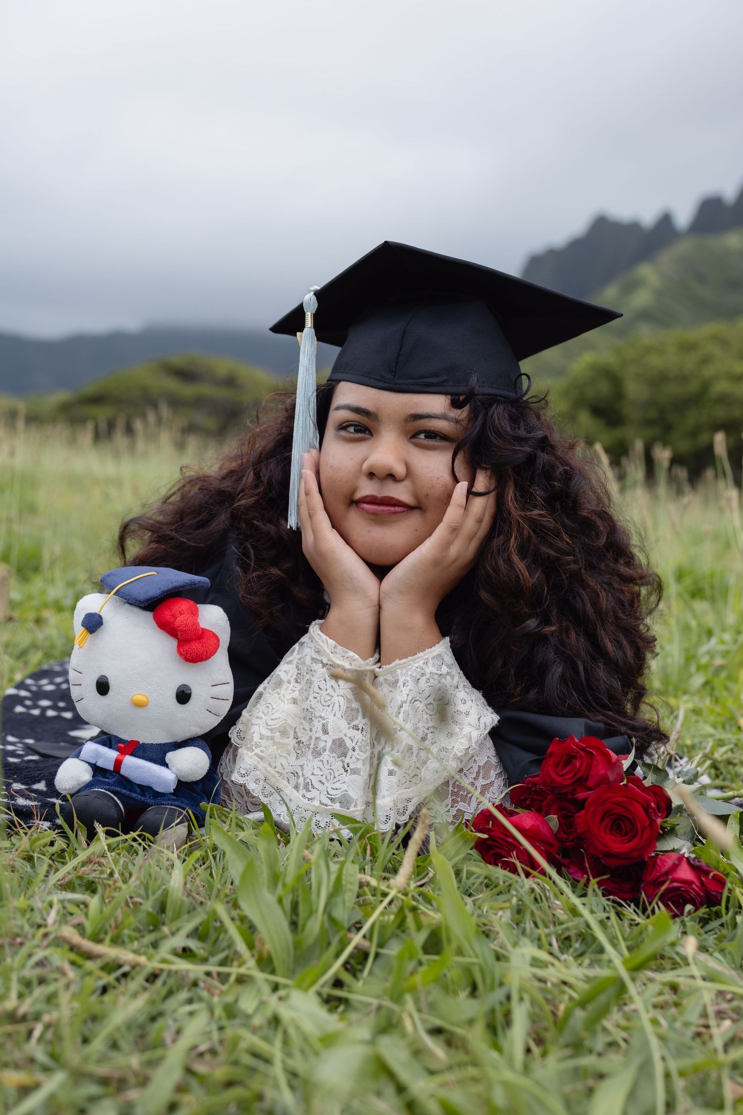 Graduate in cap and gown poses with Hello Kitty plush and red roses on green grass with mountains behind.