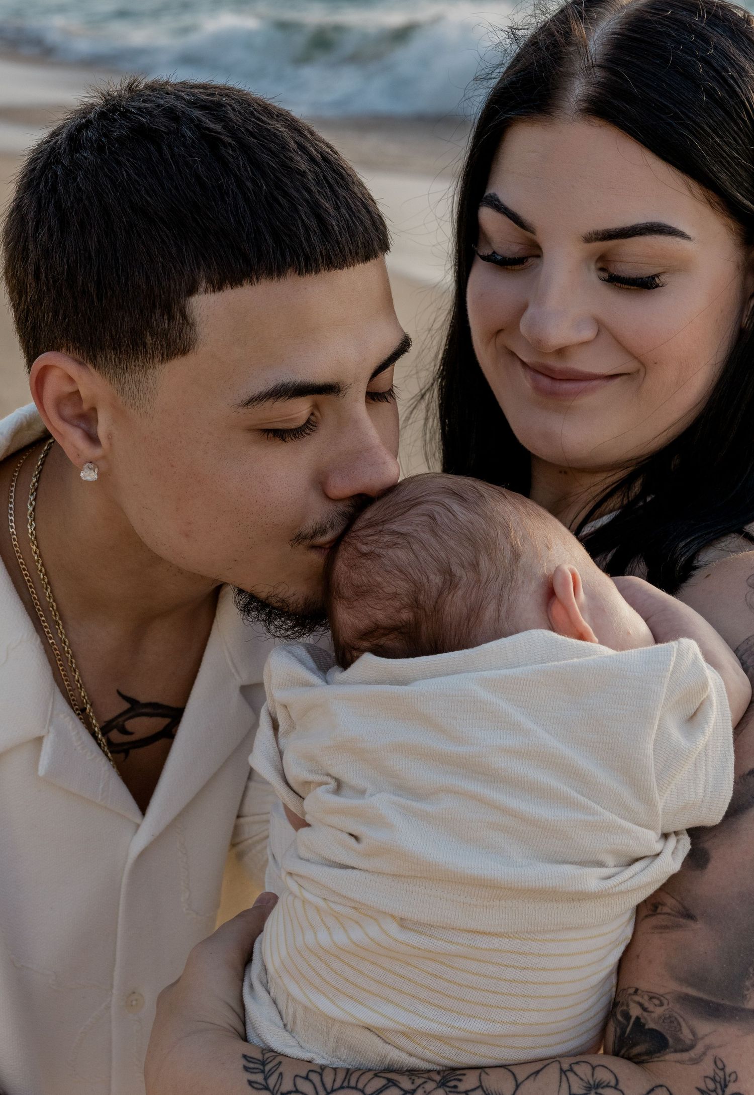 Father kissing newborn baby's head while tattooed mother watches lovingly on beach.