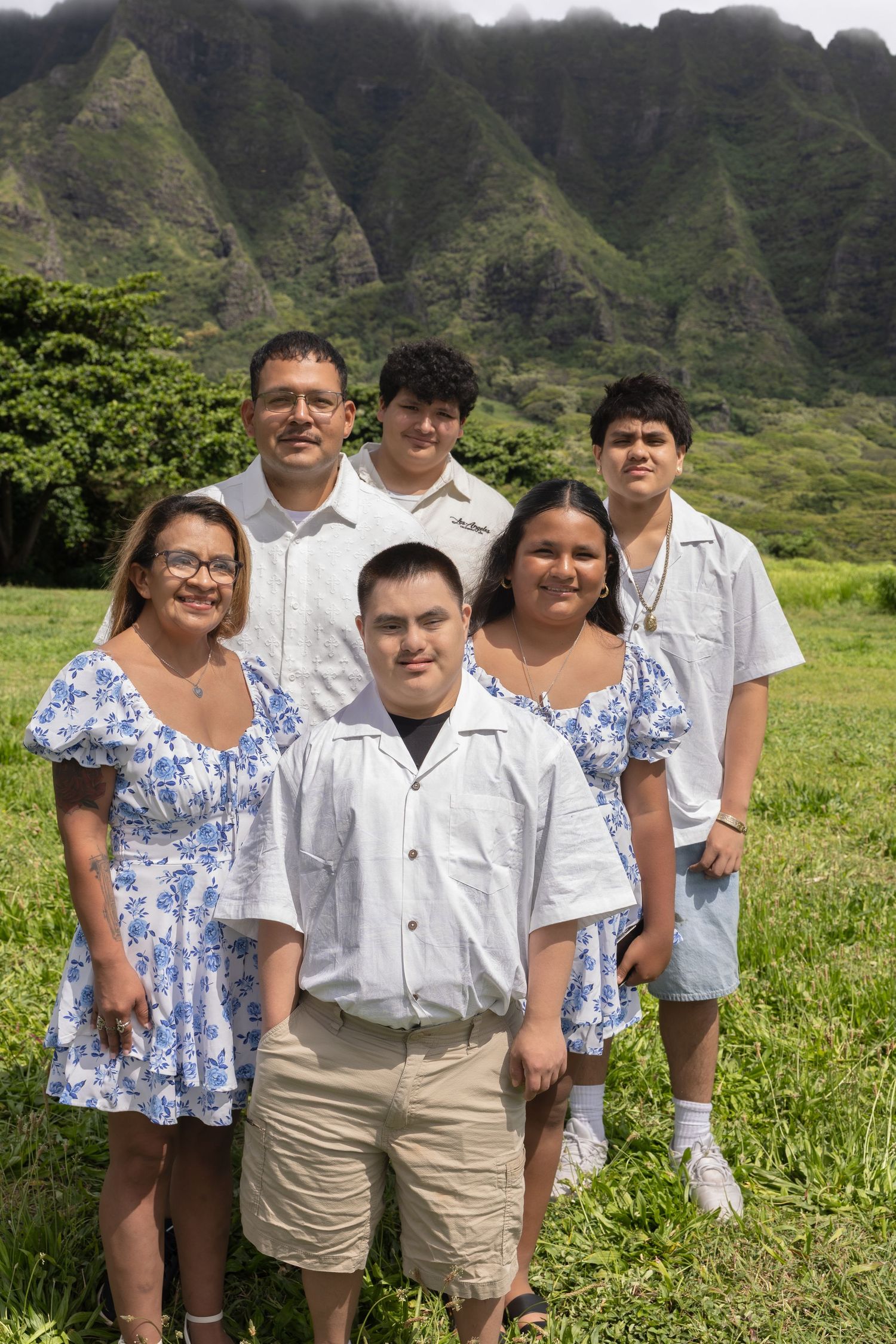 Happy family of six posing in coordinating blue floral and white outfits with lush Hawaii mountains behind.