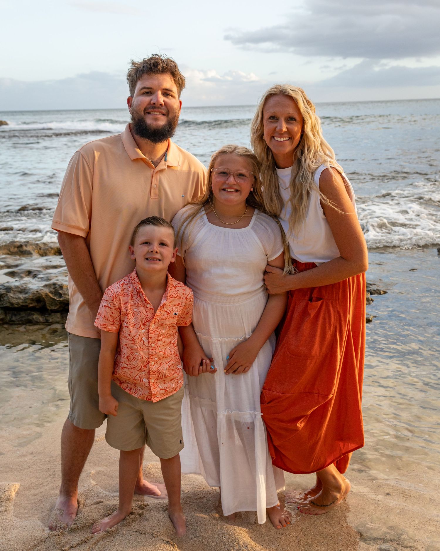 Family of four smiling together on a sandy beach at sunset, wearing coordinated outfits in warm tones.