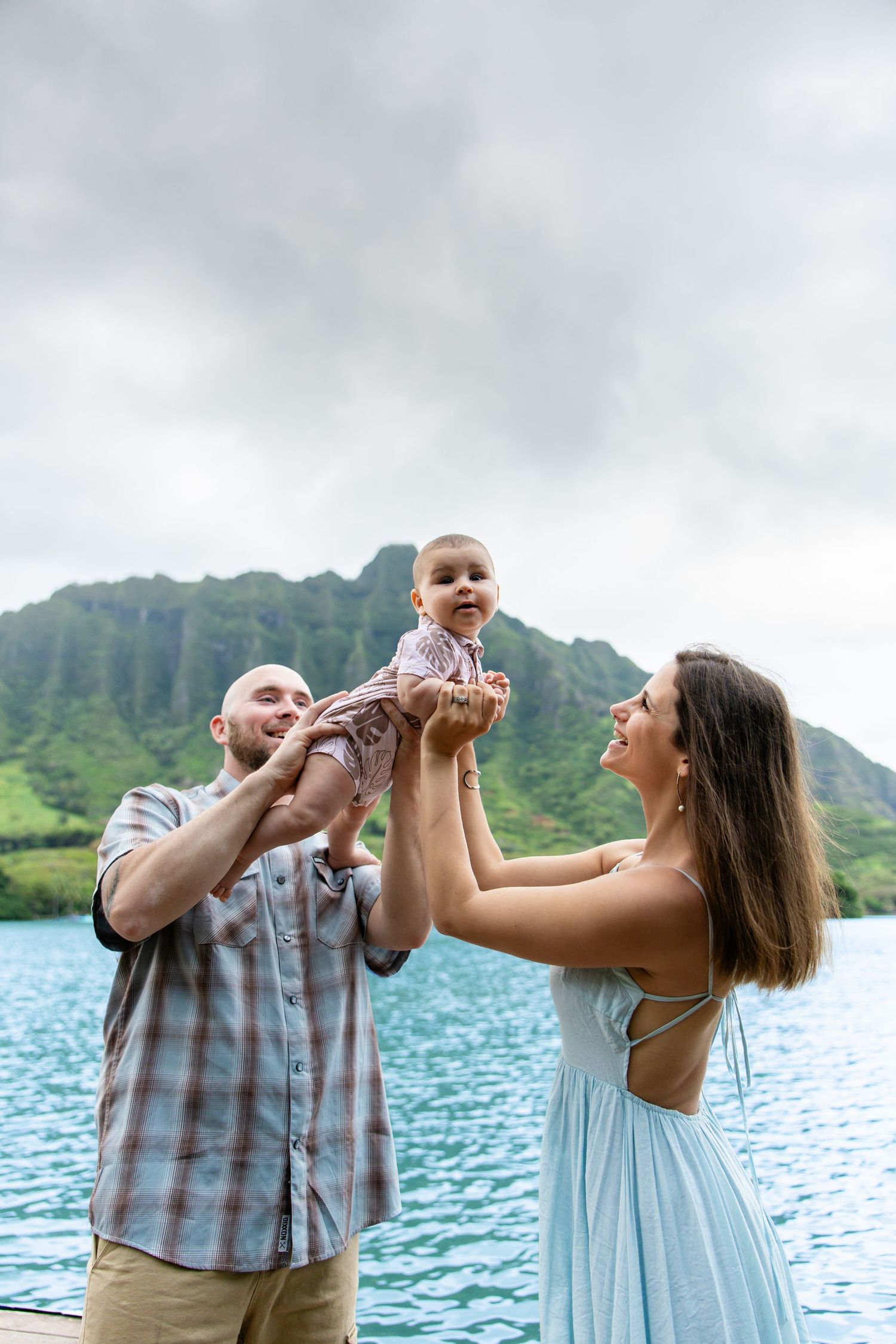 Family photo with parents lifting baby near turquoise water with green mountains in background.