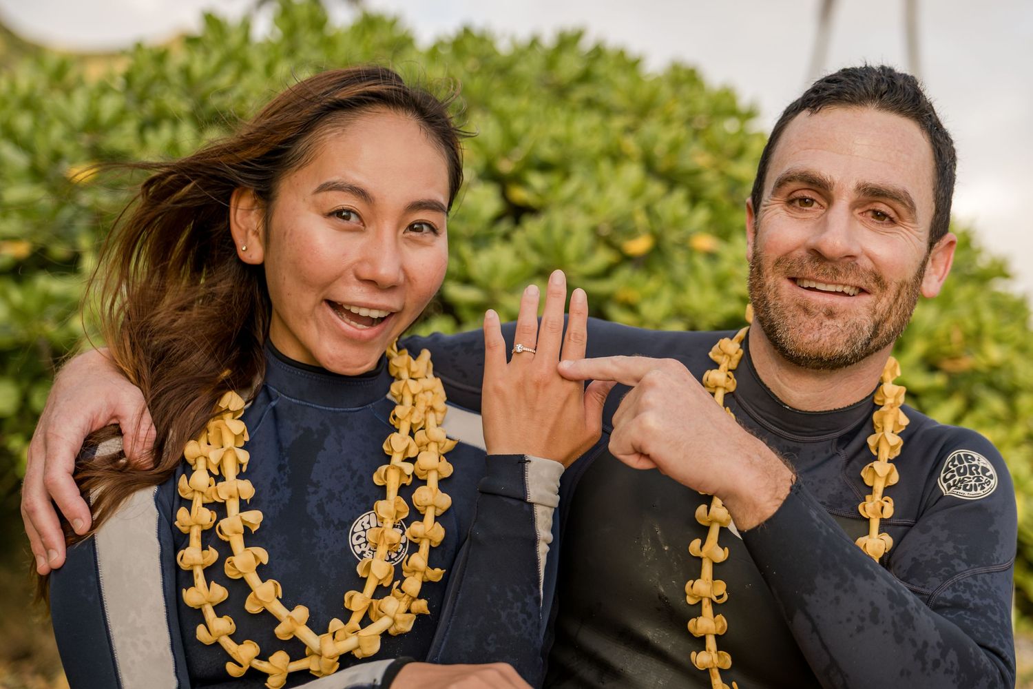 Couple wearing leis and wetsuits smiling and showing off engagement ring outdoors.