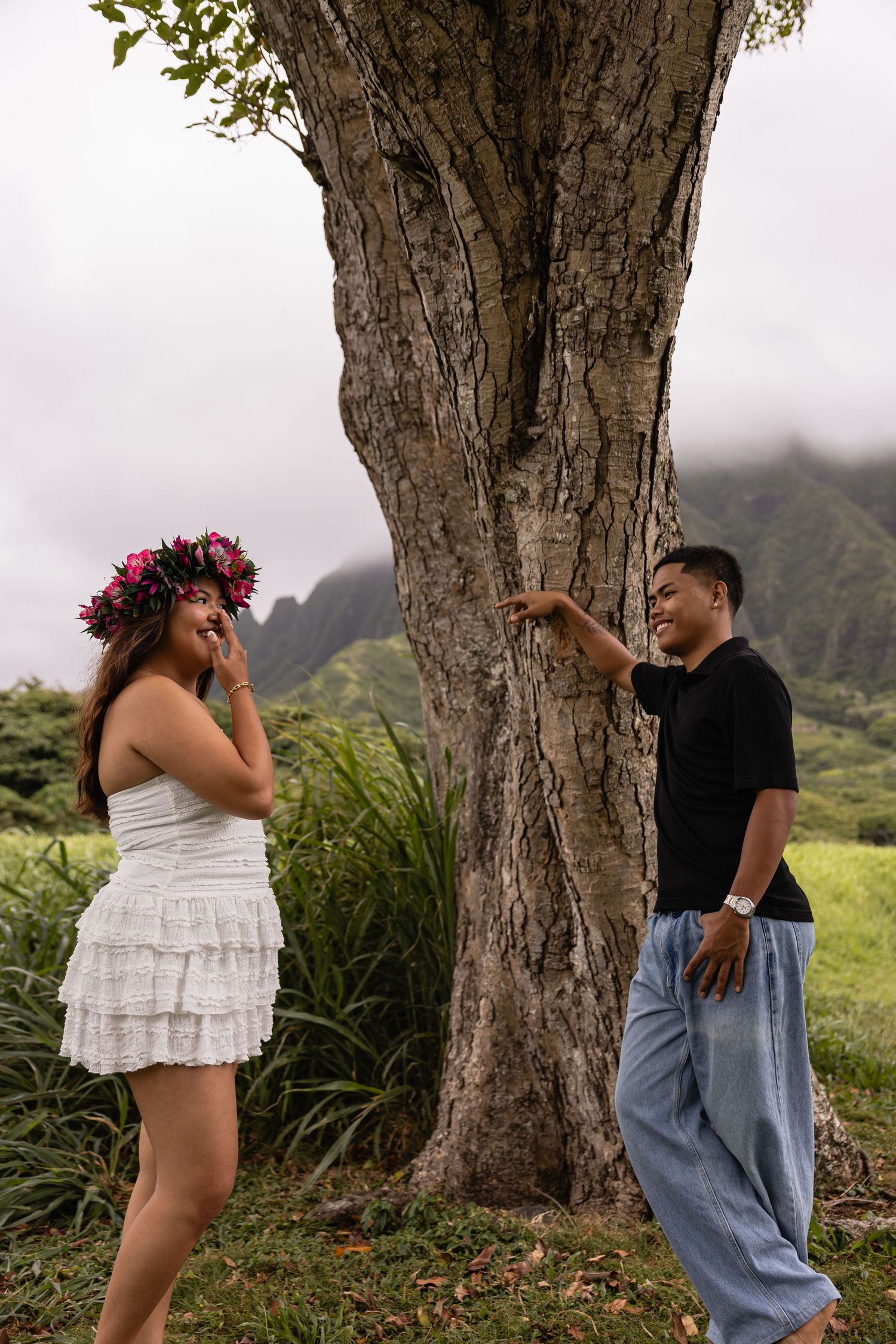 Couple laughing by a large tree in Hawaii, woman wearing floral crown and white dress, man in black shirt and blue jeans.