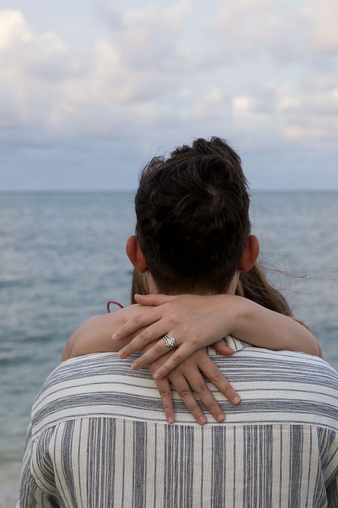 Close-up of woman's hand with engagement ring wrapped around man's shoulders at the beach.
