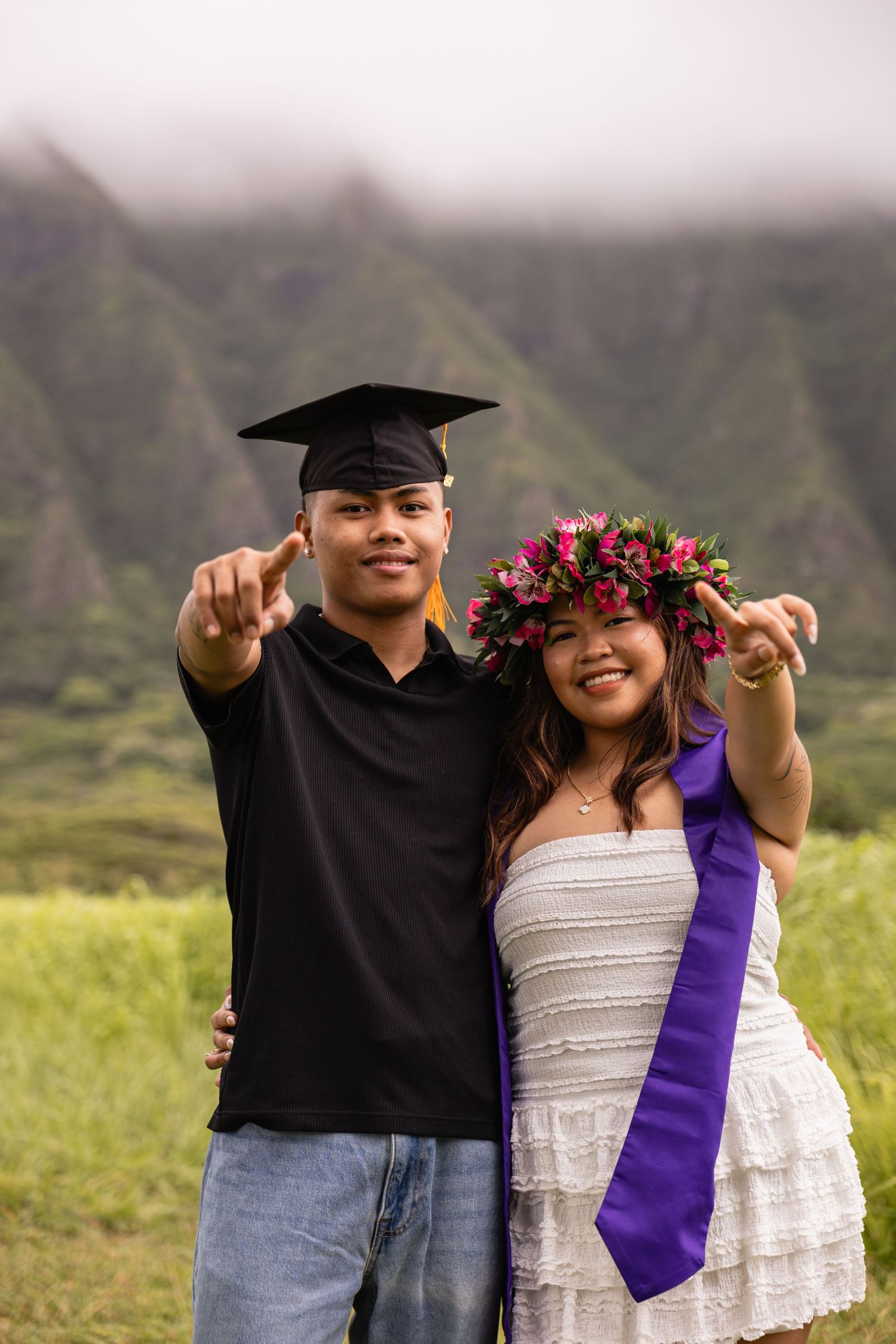 Graduate in cap and gown with woman in floral lei crown pointing at camera, lush Hawaiian mountains in background.