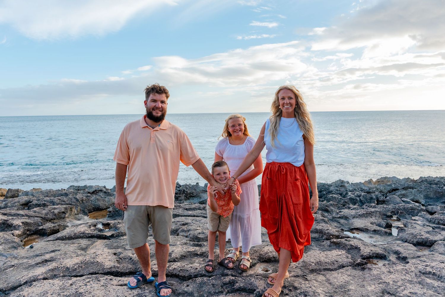 Happy family of four posing on rocky shoreline with ocean and cloudy sky backdrop during beach vacation.