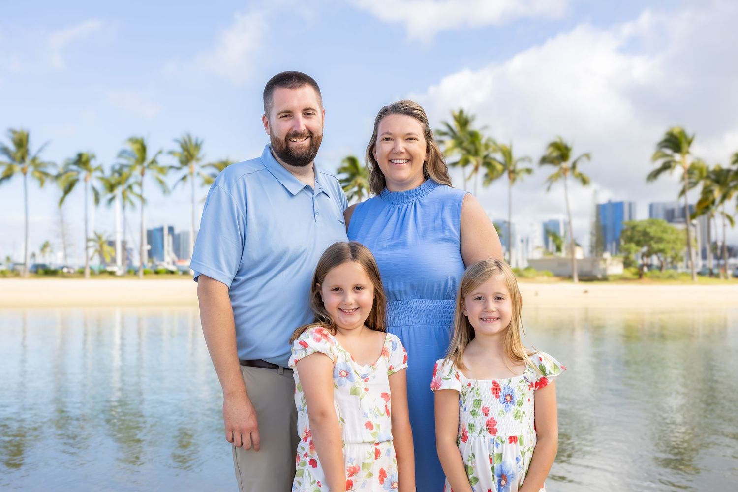 Family of four smiling near a tropical beach with palm trees and a city skyline in the background.