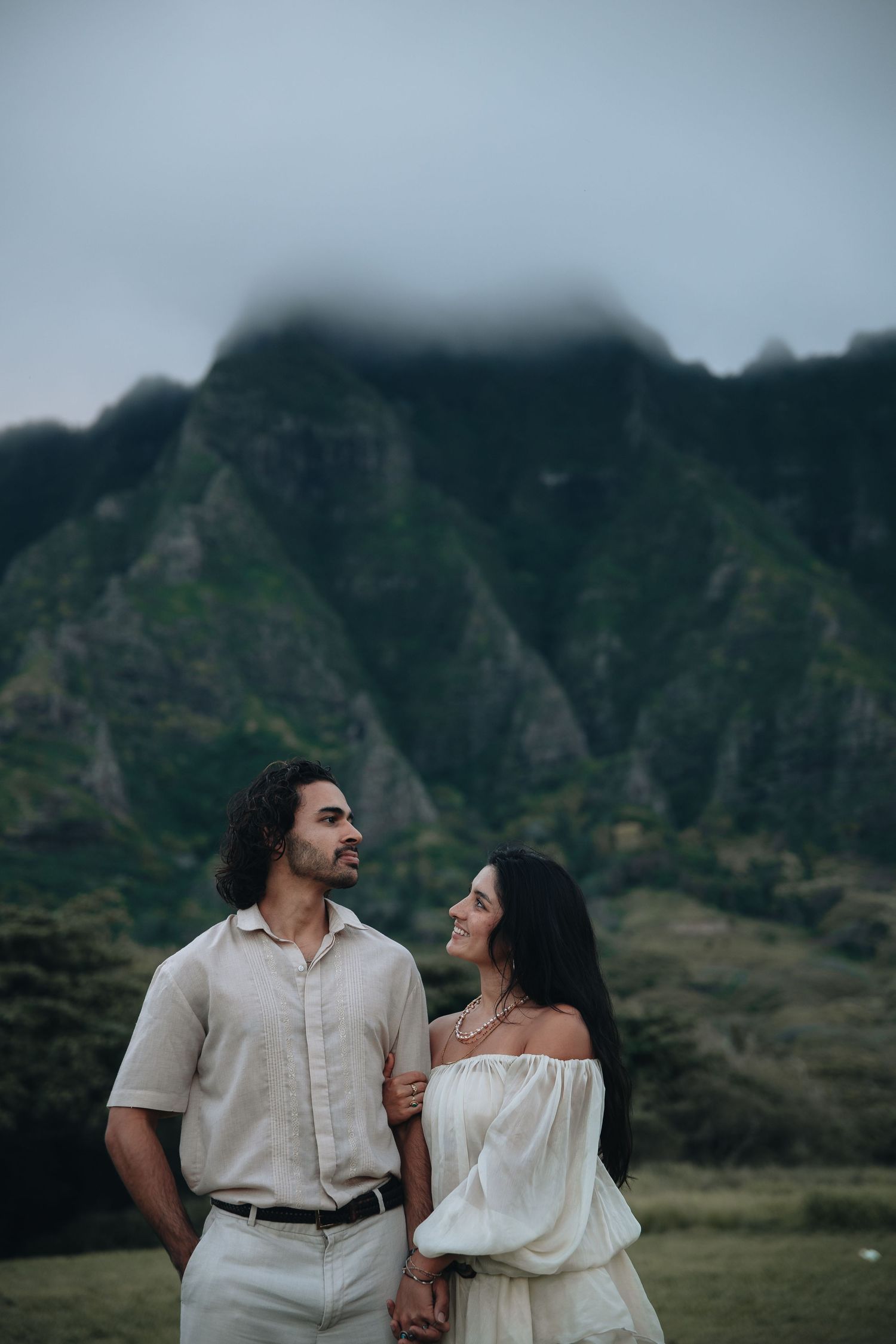 Couple holding hands and gazing at each other with dramatic cloud-covered Kualoa mountains behind.