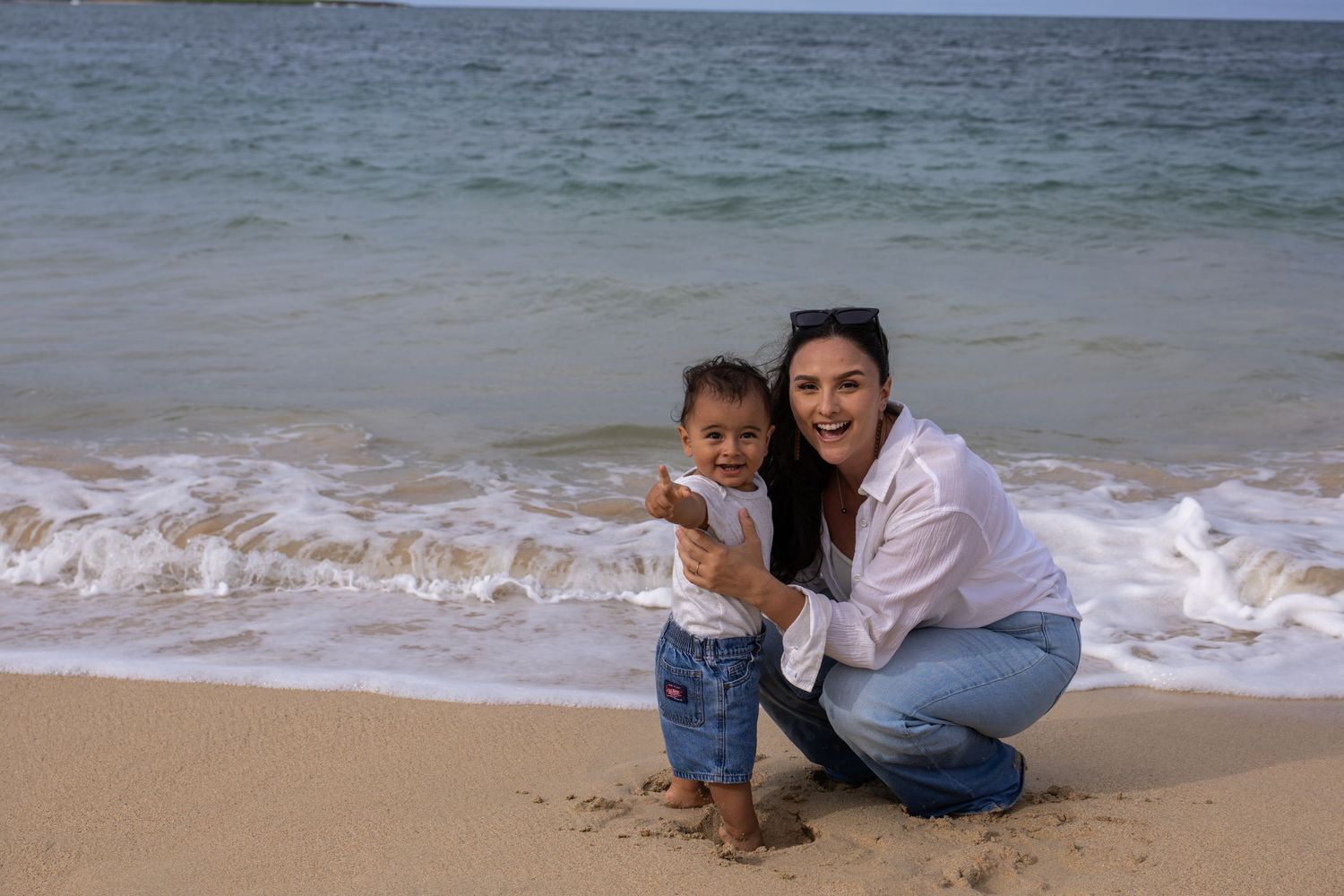 Smiling woman crouching beside standing toddler on sandy beach with ocean waves approaching shore.