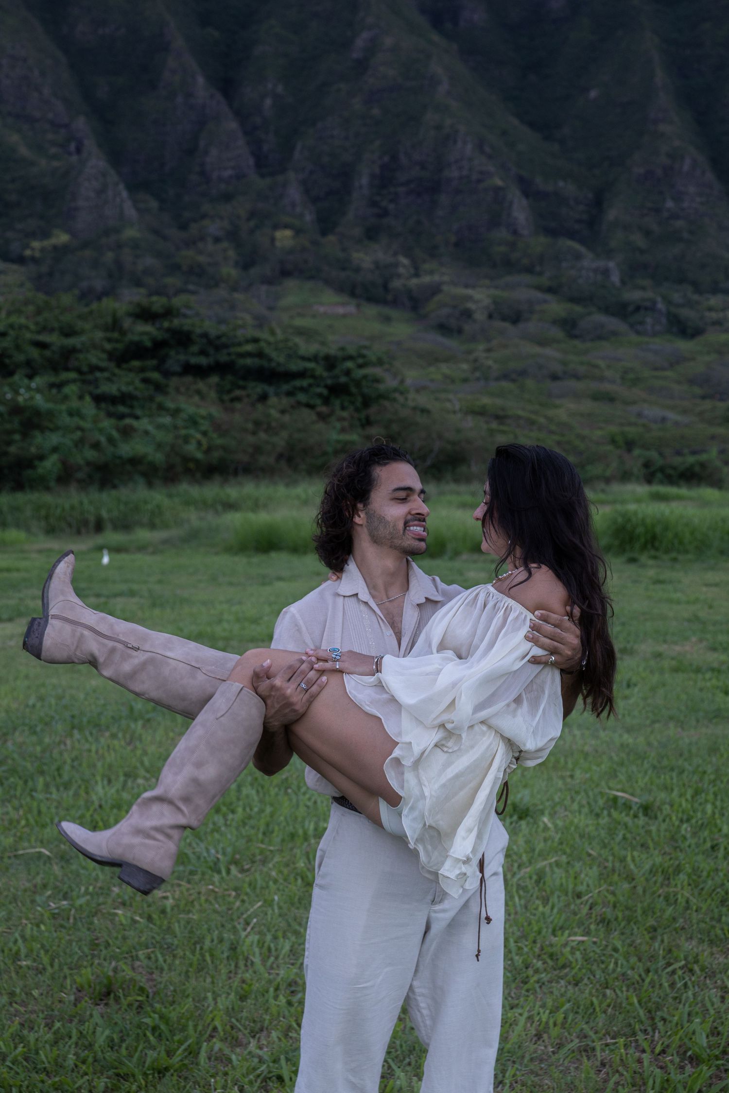 Man lifting woman in white dress and tan boots in front of dramatic green Hawaii mountain landscape.