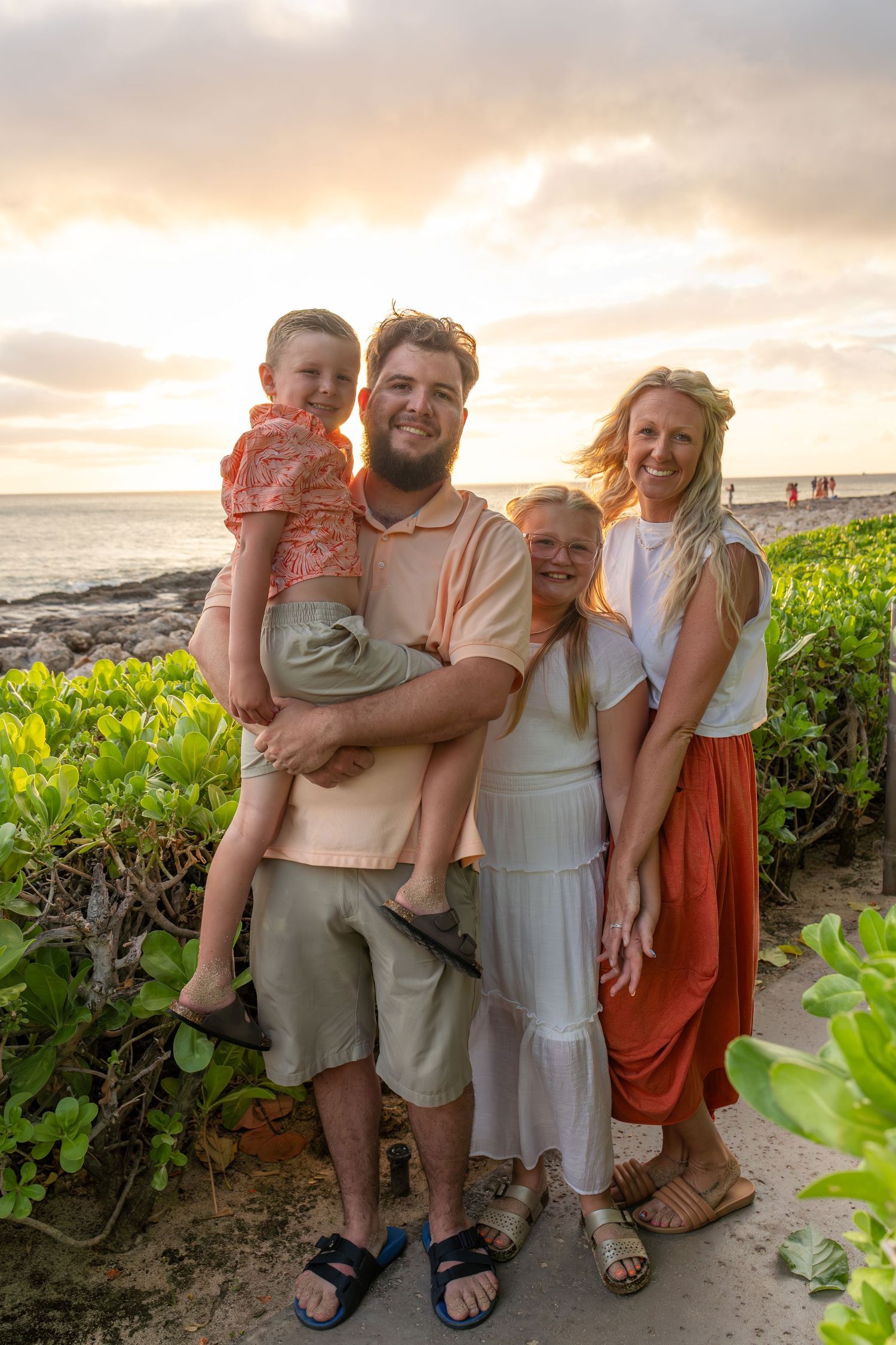 Family of four smiling at sunset on a tropical beach, man holding young child, women in white and orange dresses.