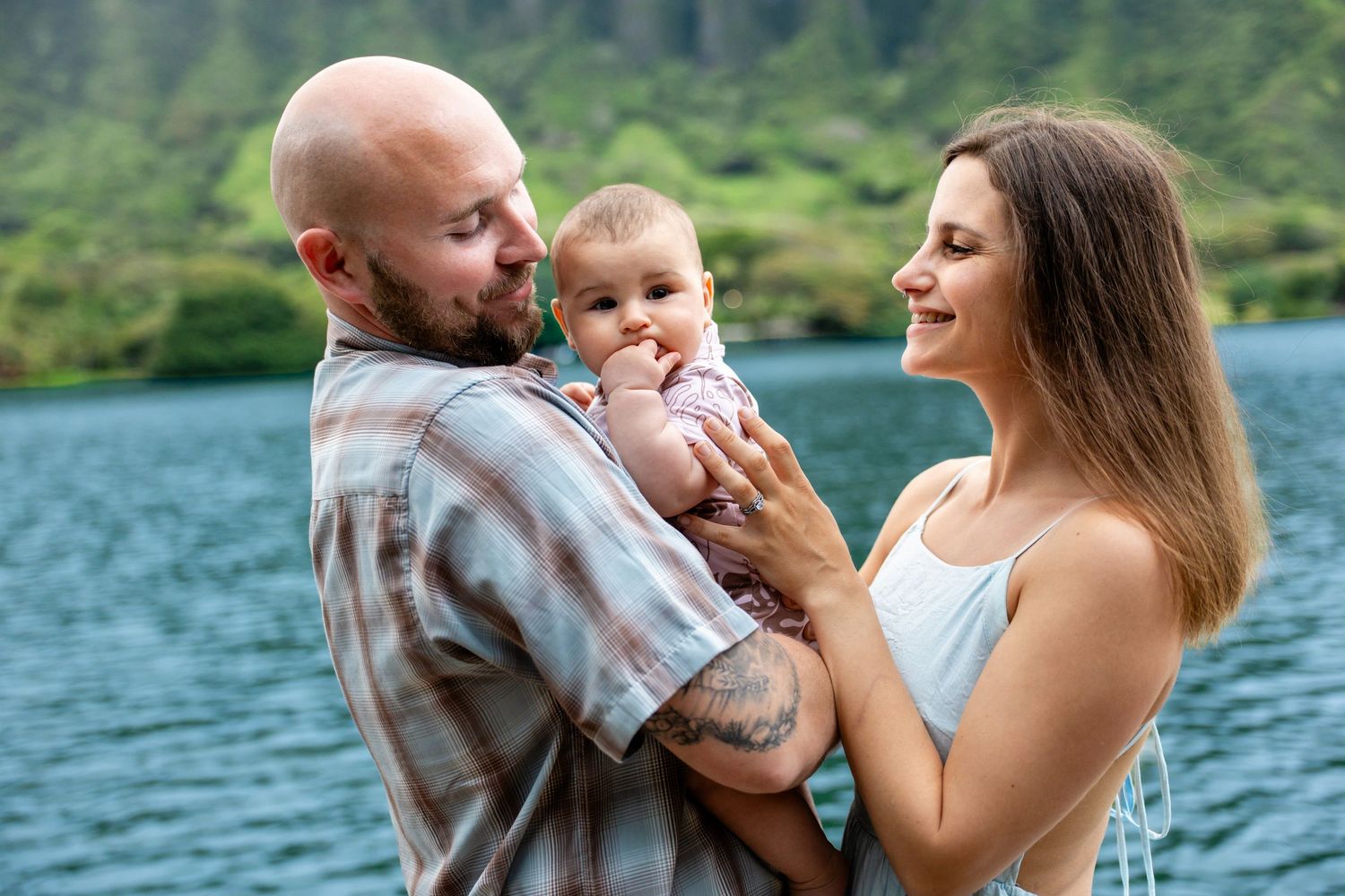 A happy young family with a bald tattooed father and brunette mother holding their baby by a scenic mountain lake.