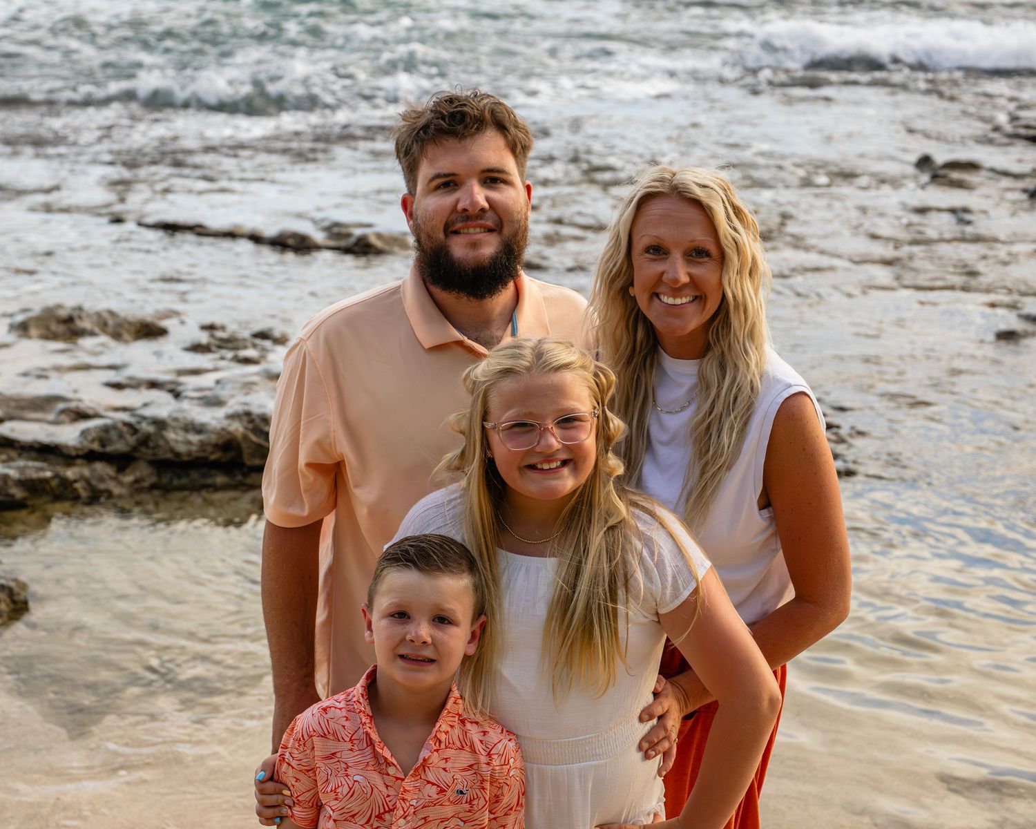 Family of four smiling together on a rocky beach, parents with two children enjoying a sunny coastal vacation.