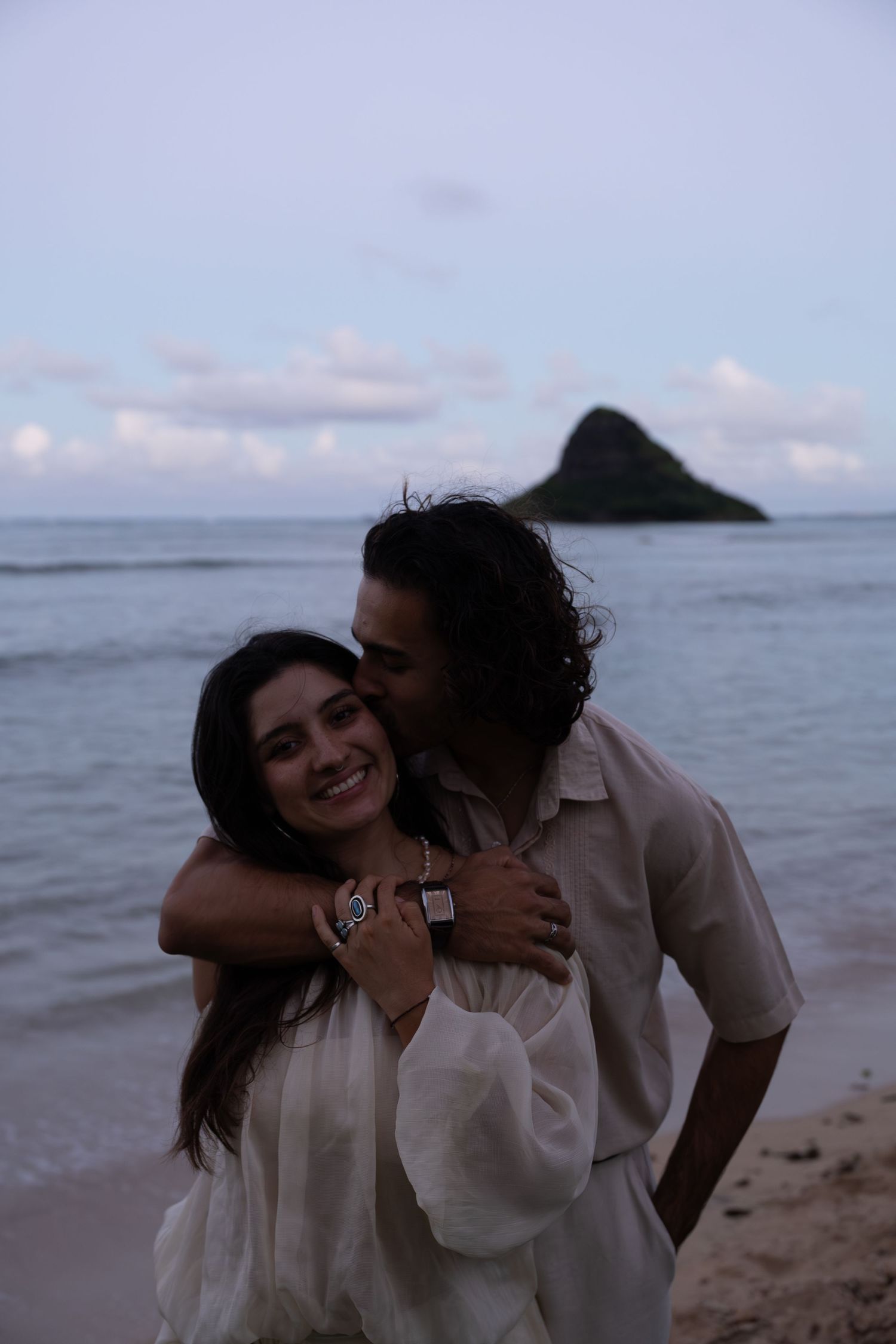 A couple embraces on a Hawaiian beach at dusk, with Mokoliʻi Island visible in the calm ocean behind them.