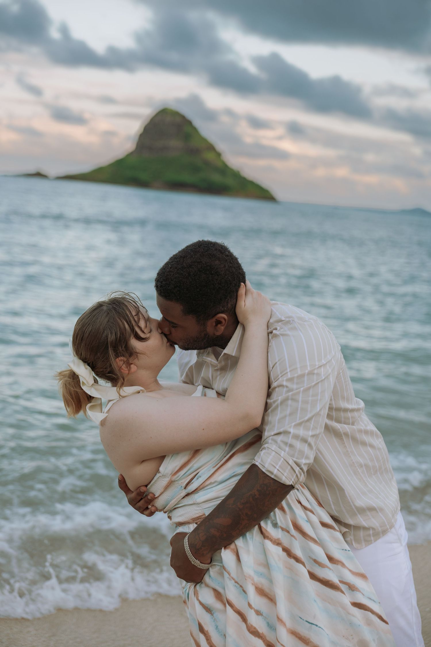 Interracial couple sharing a passionate kiss on a Hawaiian beach with Chinaman's Hat island backdrop.