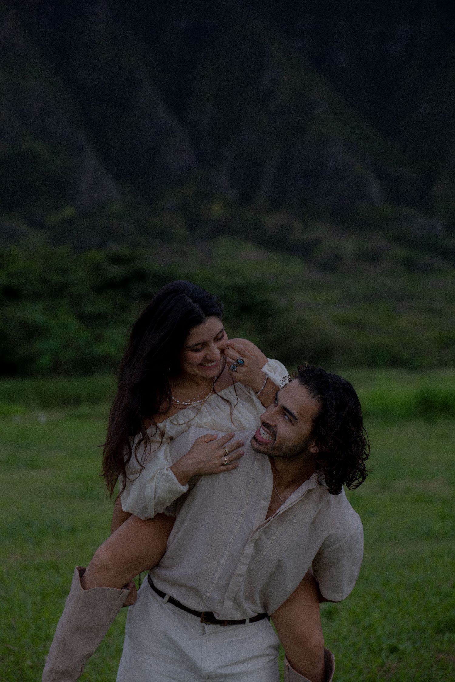 Man giving woman piggyback ride laughing in front of lush green mountain landscape.