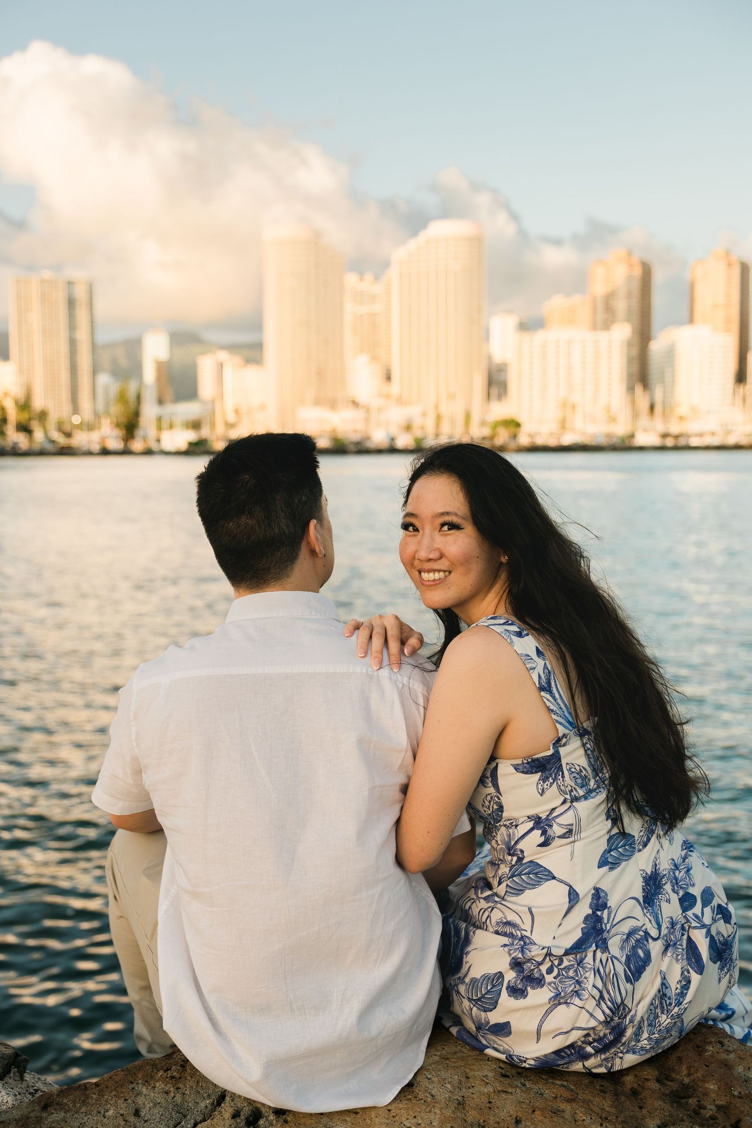 Woman in floral dress smiles over shoulder while sitting with partner against Honolulu city skyline.