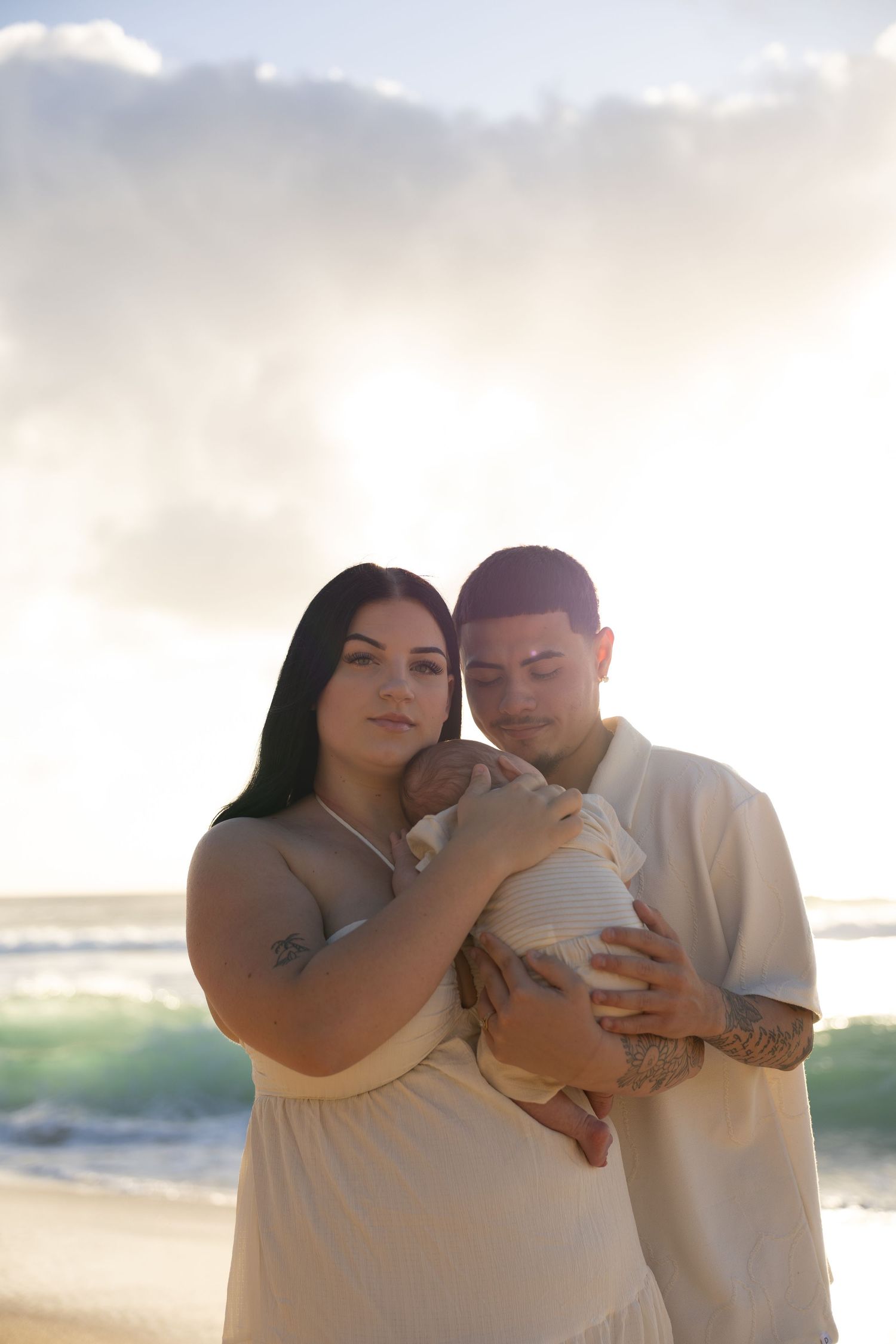 Young couple holding newborn baby on beach at sunset, wearing cream outfits with ocean waves behind them.