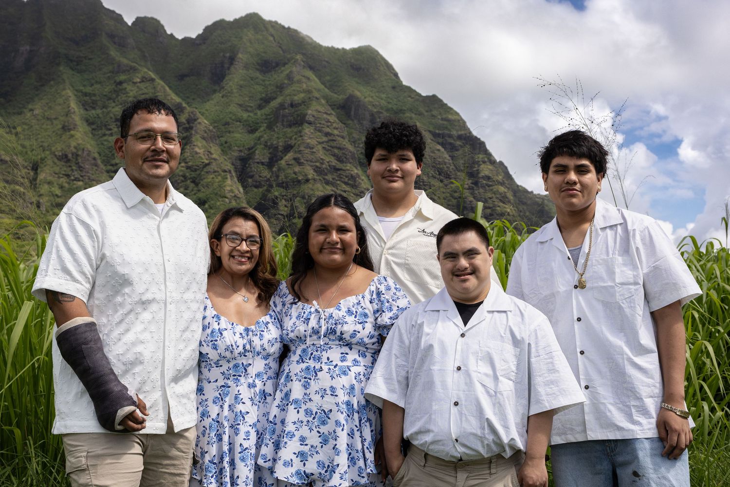 Happy family of six smiling together outdoors with stunning green Hawaiian mountain range backdrop.