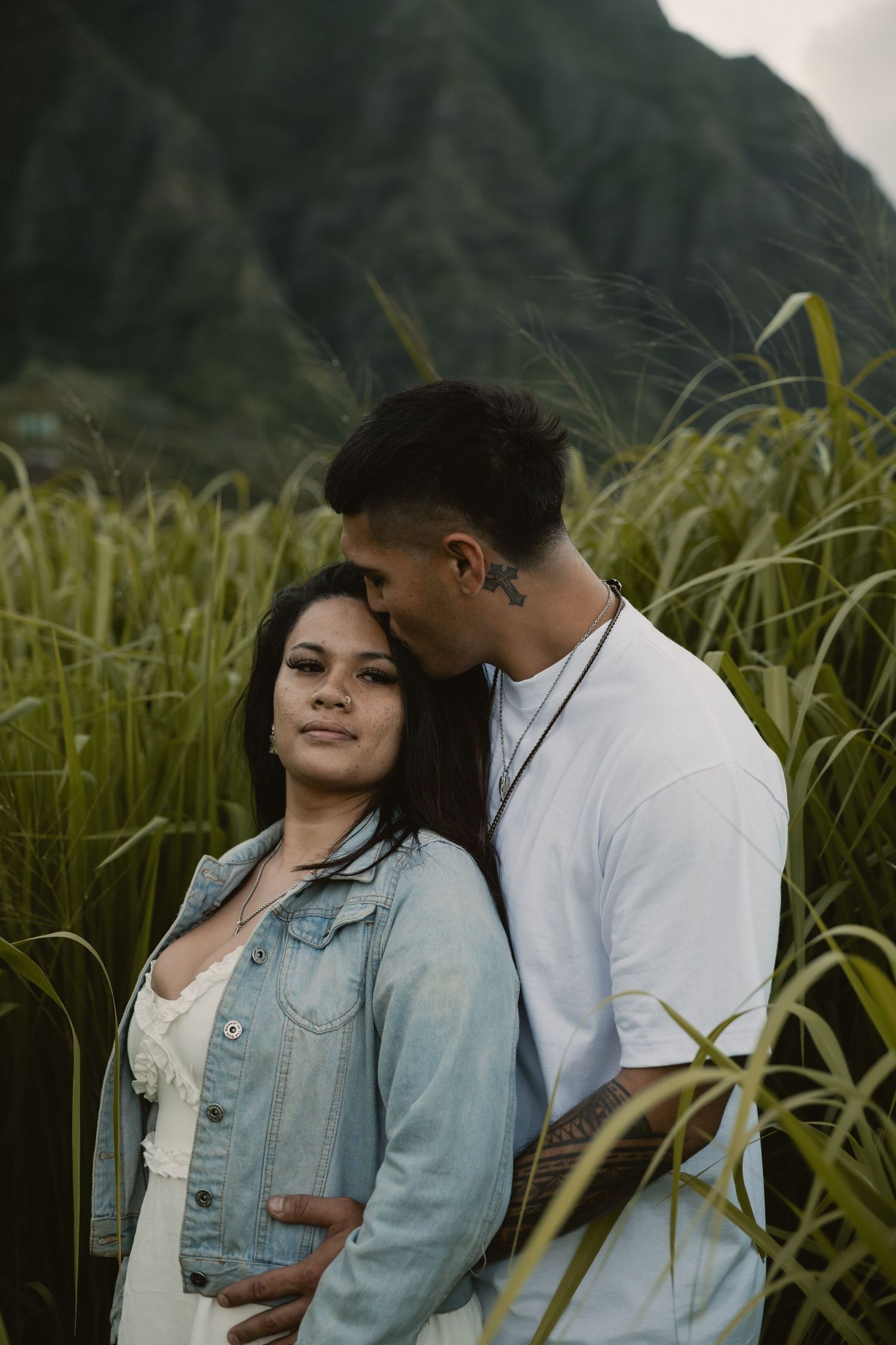 A couple embraces in a lush green field with misty mountains, woman in denim jacket smiles softly as man kisses her head.