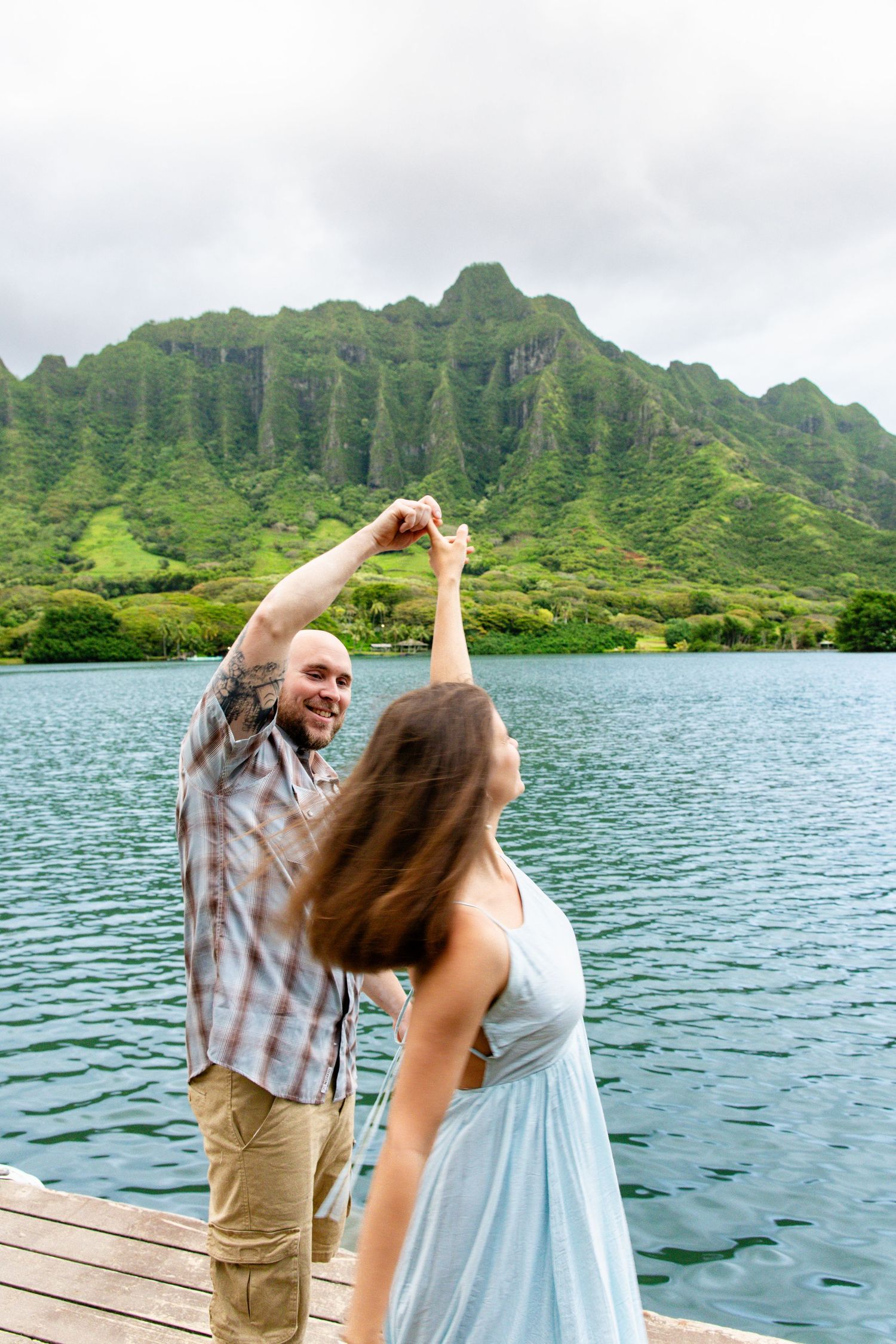Couple dancing on a wooden dock with lush green mountains and turquoise lake in Hawaii background.
