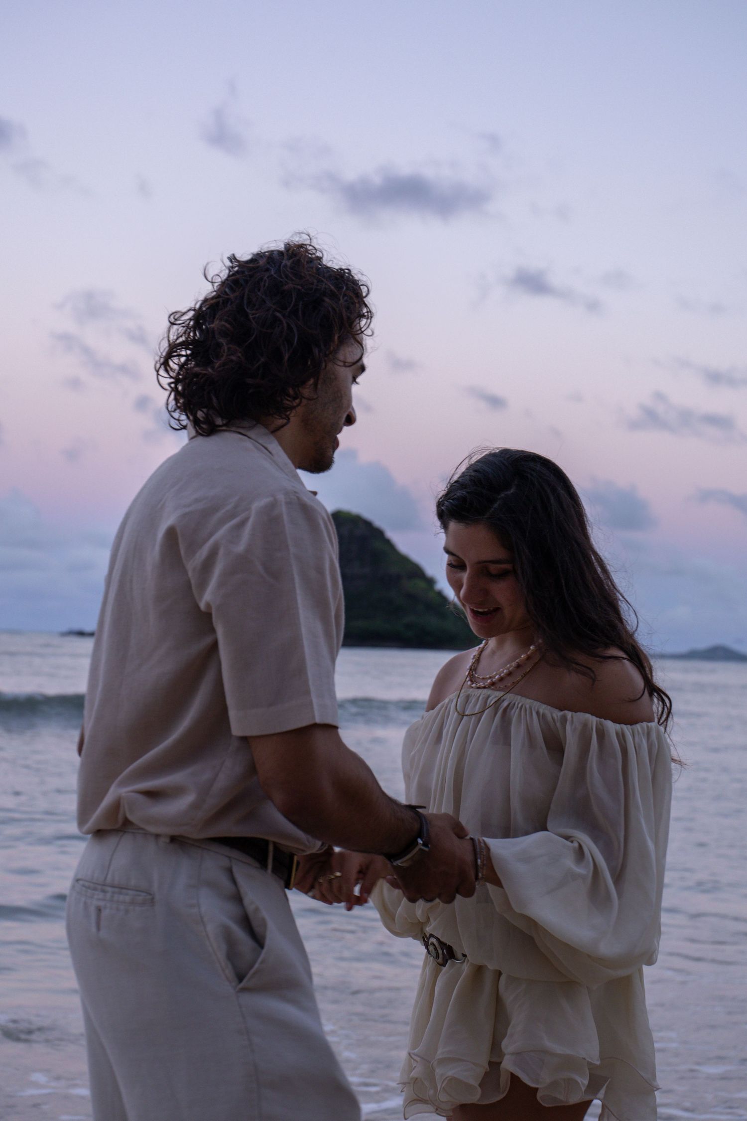 Couple holding hands on beach at sunset with island silhouette and pink sky behind.