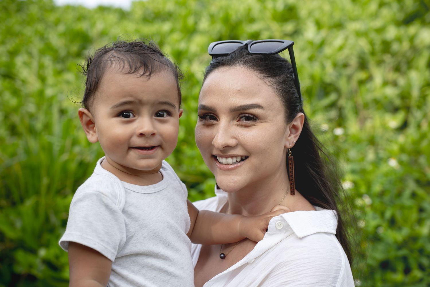 Beautiful woman holds smiling baby boy outdoors with green foliage background.