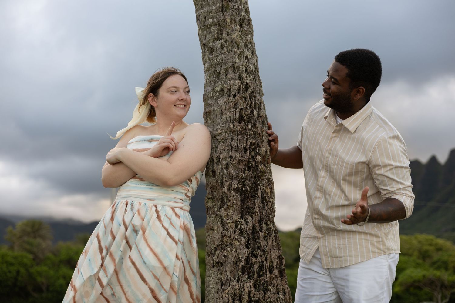 Smiling couple laughing together beside a tree trunk with cloudy mountain scenery in Hawaii.
