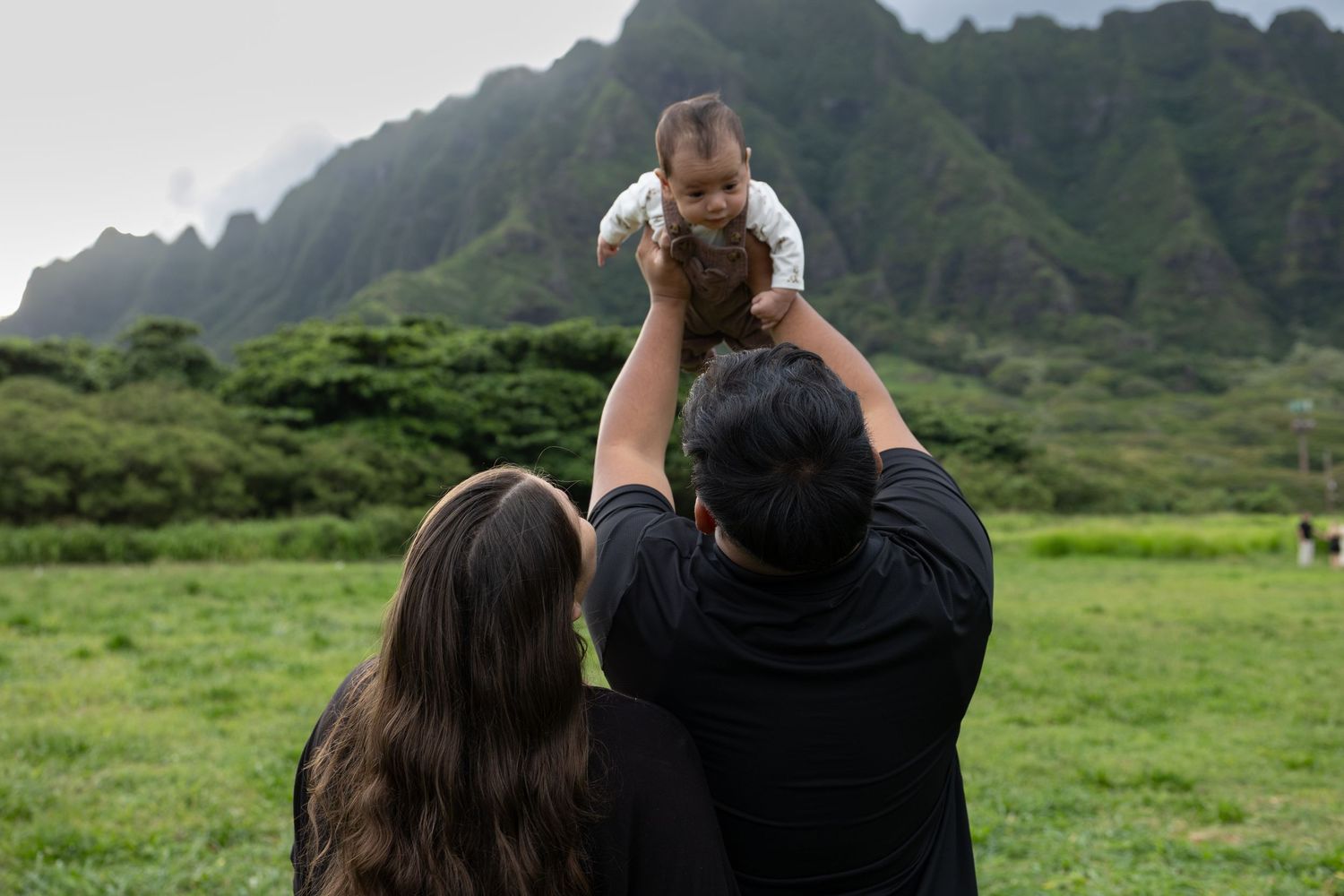 Parents lifting baby in the air with lush green mountains in Hawaii background.
