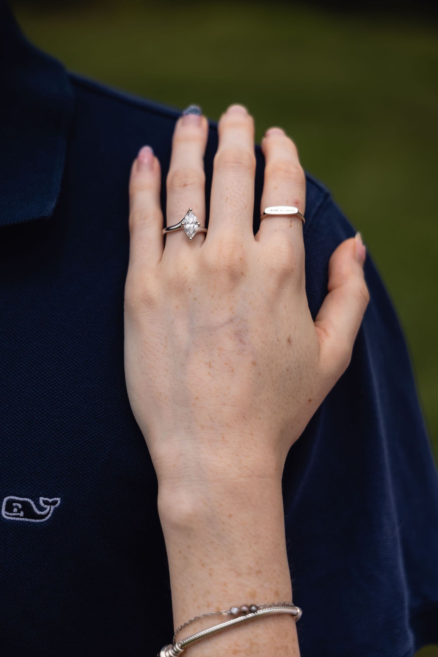 Close-up of a woman's hand wearing a diamond engagement ring and gold band, with silver bracelets, on a navy shirt.