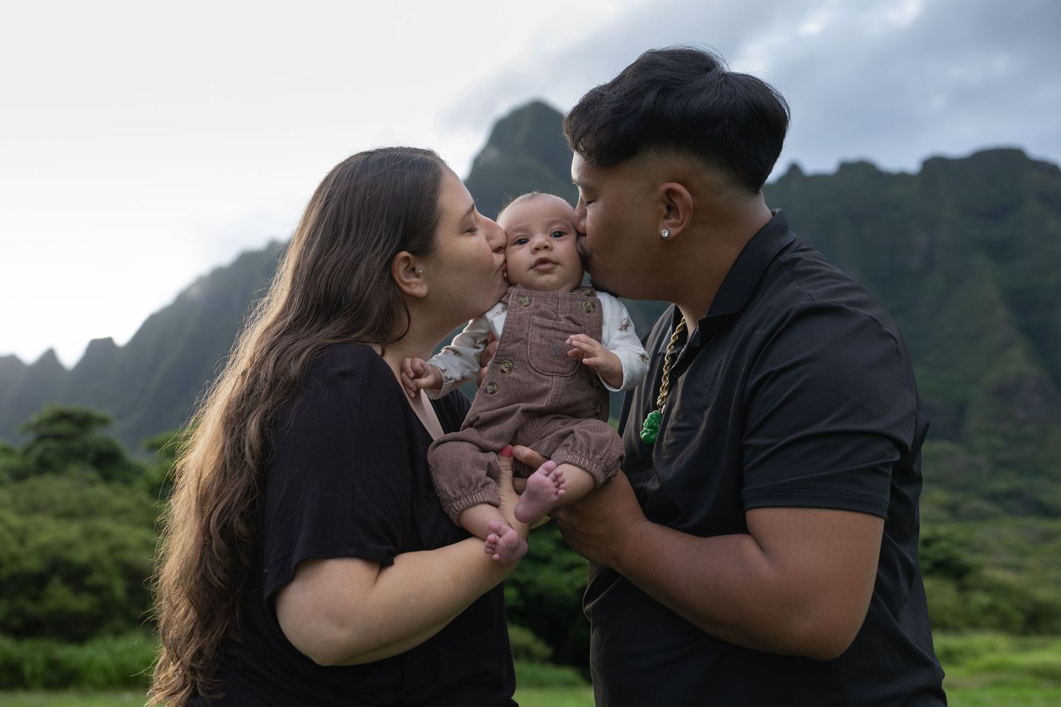 Parents kissing their baby on the cheeks with Hawaii mountains in background.