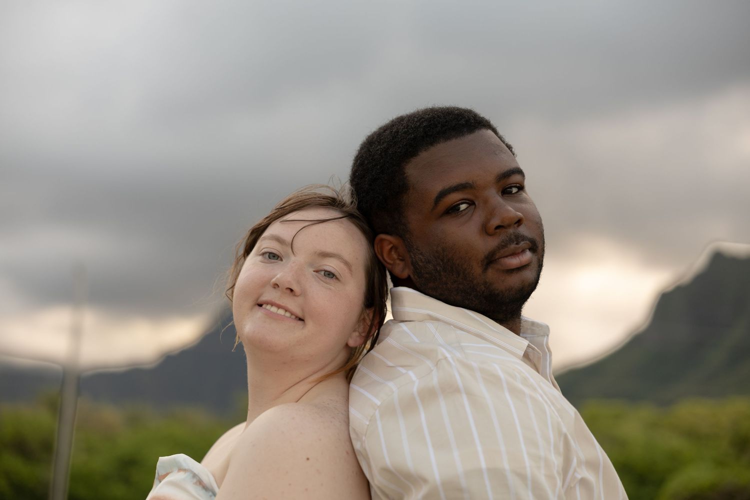 Couple posing back to back smiling with dramatic green Hawaiian mountains in the background.